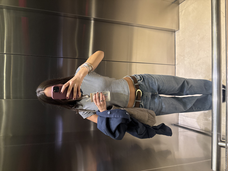 An individual, appearing to be female with long dark hair, is depicted taking a mirror selfie inside an elevator. Her face is obscured by the smartphone she holds in her right hand, which has a purple protective case and dark red painted fingernails. She wears a fitted black and white horizontally striped crop top, revealing a portion of her abdomen, and light blue relaxed-fit jeans with a frayed waistband, secured by a dark brown belt with a gold buckle. Her left wrist displays multiple silver bangles, and her left hand holds a clear glass beverage container with a straw, also featuring dark red painted nails. A navy blue jacket and a light brown quilted bag are draped over her left arm. The elevator interior features brushed stainless steel walls with vertical seams and a light-colored, possibly marble or tiled, floor. A reflective metal handrail is visible at the bottom of the frame. The scene is located within an elevator in Mexico City, Mexico.