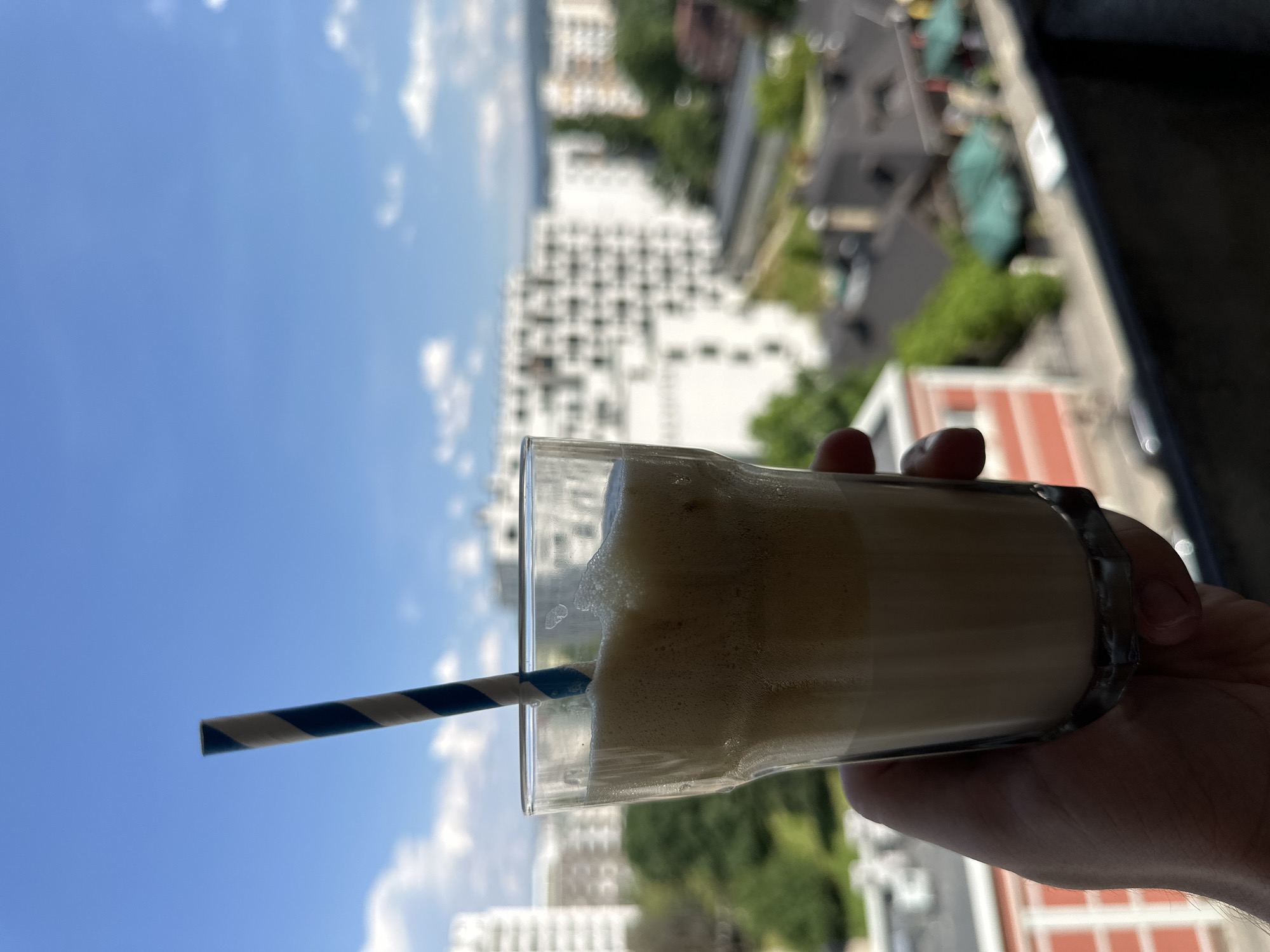 A hand holds a glass of frothy beverage with a blue and white striped straw against a backdrop of urban buildings and a blue sky. The glass appears to be filled with a drink that has a creamy, layered appearance, with foam at the top. The background shows apartment buildings, trees, and a glimpse of what looks like a balcony or rooftop railing in the foreground. The city in the background is consistent with the architecture found in Sofia, Bulgaria, particularly the style of the residential buildings and the general urban landscape. The weather appears to be clear and sunny.