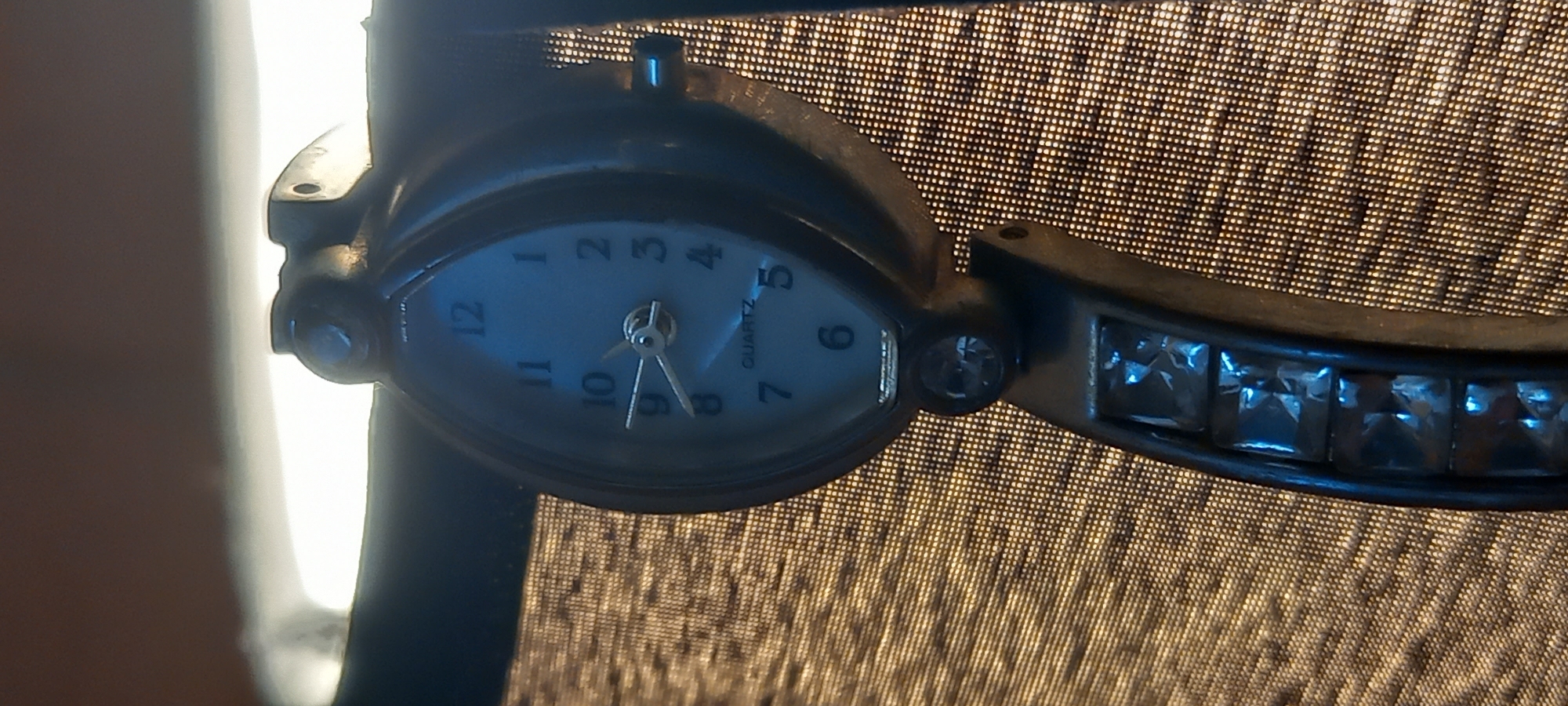 This is a close-up, vertical, eye-level shot of a wristwatch. The watch face is oval-shaped with a light blue background and black numbers. The time displayed is approximately 9:40. The watch band is made of dark metal and is adorned with square, clear, faceted stones. The watch is hanging on a dark fabric, possibly a curtain or upholstery, which has a textured, woven appearance and is illuminated from behind, creating a bokeh effect. The watch and the fabric are the sole subjects in the image. There are no people or any indication of the city of Kryvyi Rih, Ukraine.