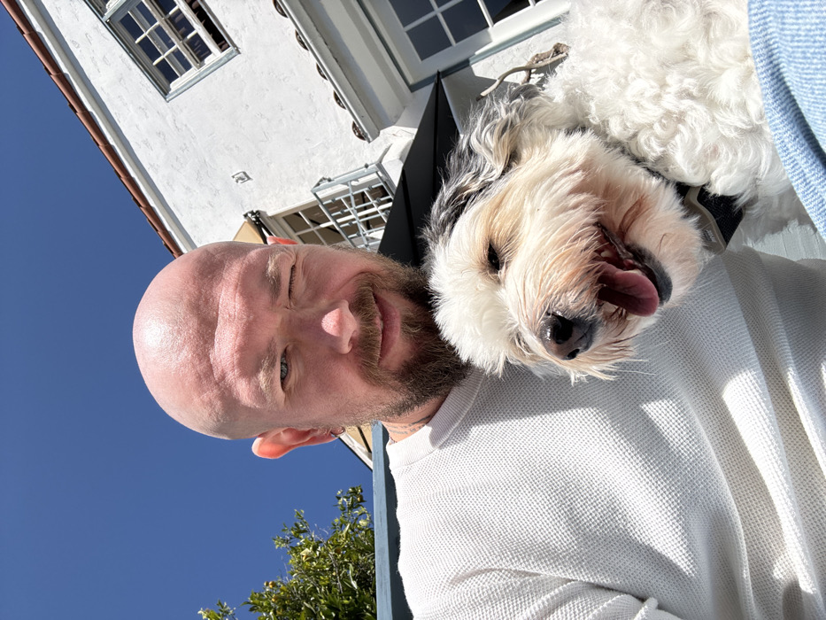 The image displays an outdoor selfie taken in Los Angeles, United States of America, featuring one adult male and one domestic animal. The male subject, bald with a light-colored beard and mustache, wears a white waffle-knit long-sleeved shirt. He is winking with his right eye and smiling directly at the camera. Cradled against his chest is a small dog, appearing to be a shaggy white and black terrier-type mix, with its mouth open, tongue slightly extended, and eyes partially closed. The dog is wearing a dark-colored collar. The background consists of a clear blue sky on the left and a white stucco building with a red tile roof on the right. The building features multiple windows with white frames, a small balcony with a railing, and a dark awning. Green foliage is visible in the upper left background. The scene is brightly illuminated by natural sunlight, casting distinct shadows.