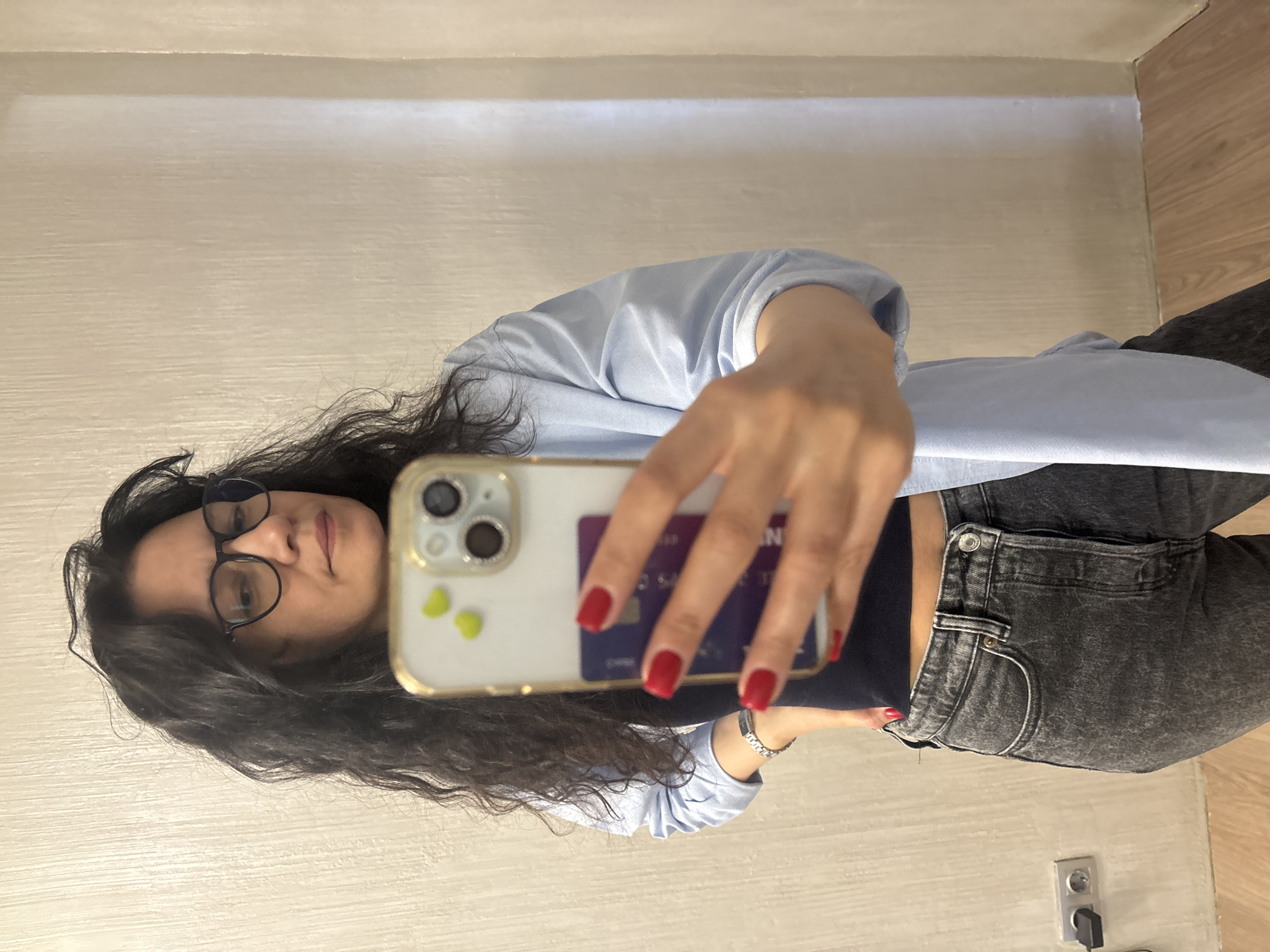 A woman takes a selfie. She is wearing glasses, a light blue button-down shirt, and dark gray jeans. Her fingernails are painted red. She is holding a phone with a credit card tucked behind it. The background is a light-colored wall and wooden floor. There is an electrical outlet visible near her jeans. The location is Chaikovsky, Russia.