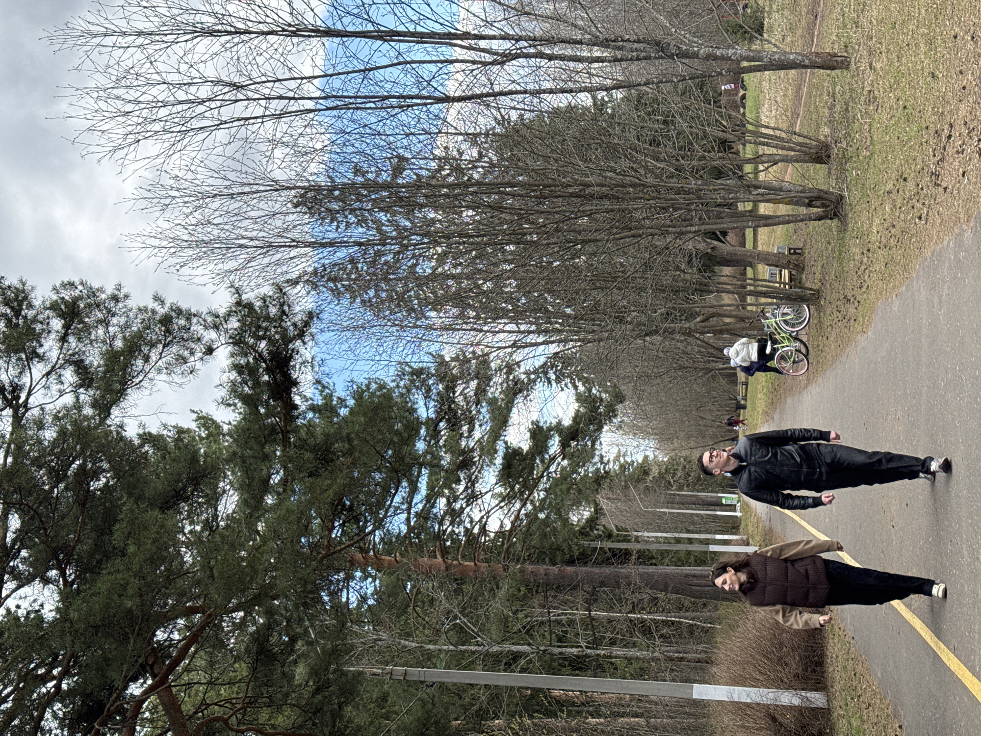 Two people are walking on a paved path through a park. The person in the foreground is wearing a brown vest and dark pants. The person behind them is wearing a black jacket and pants. The path has a yellow line painted on it. There are trees on both sides of the path, some with leaves and some without. In the distance, there are people with bicycles and benches. The location is likely a park in Izhevsk, Russia, based on the environment.