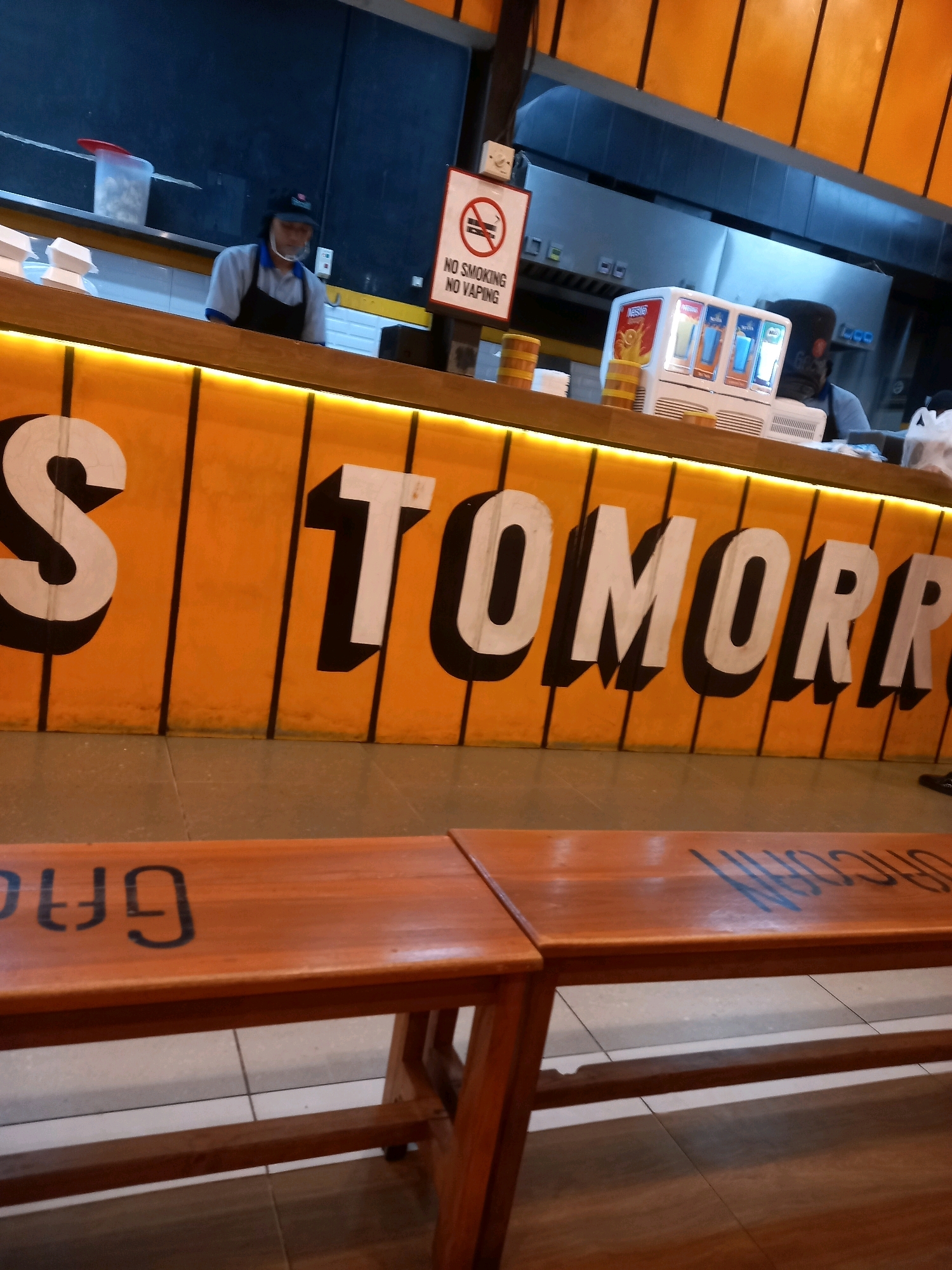 The image depicts the interior of a food establishment, possibly a fast-food restaurant, in Pamekasan, Indonesia. The most prominent feature is a brightly lit, orange counter with large white letters spelling out "TOMORROW". Behind the counter, a person wearing a dark cap, a light blue shirt, and a black apron is visible, along with a drink dispenser and kitchen equipment. A "No Smoking No Vaping" sign is also displayed. In the foreground, there are wooden benches with black lettering. One bench has the letters "UFG" visible, and the other has faded blue lettering that is difficult to decipher. The overall atmosphere suggests a casual dining setting.