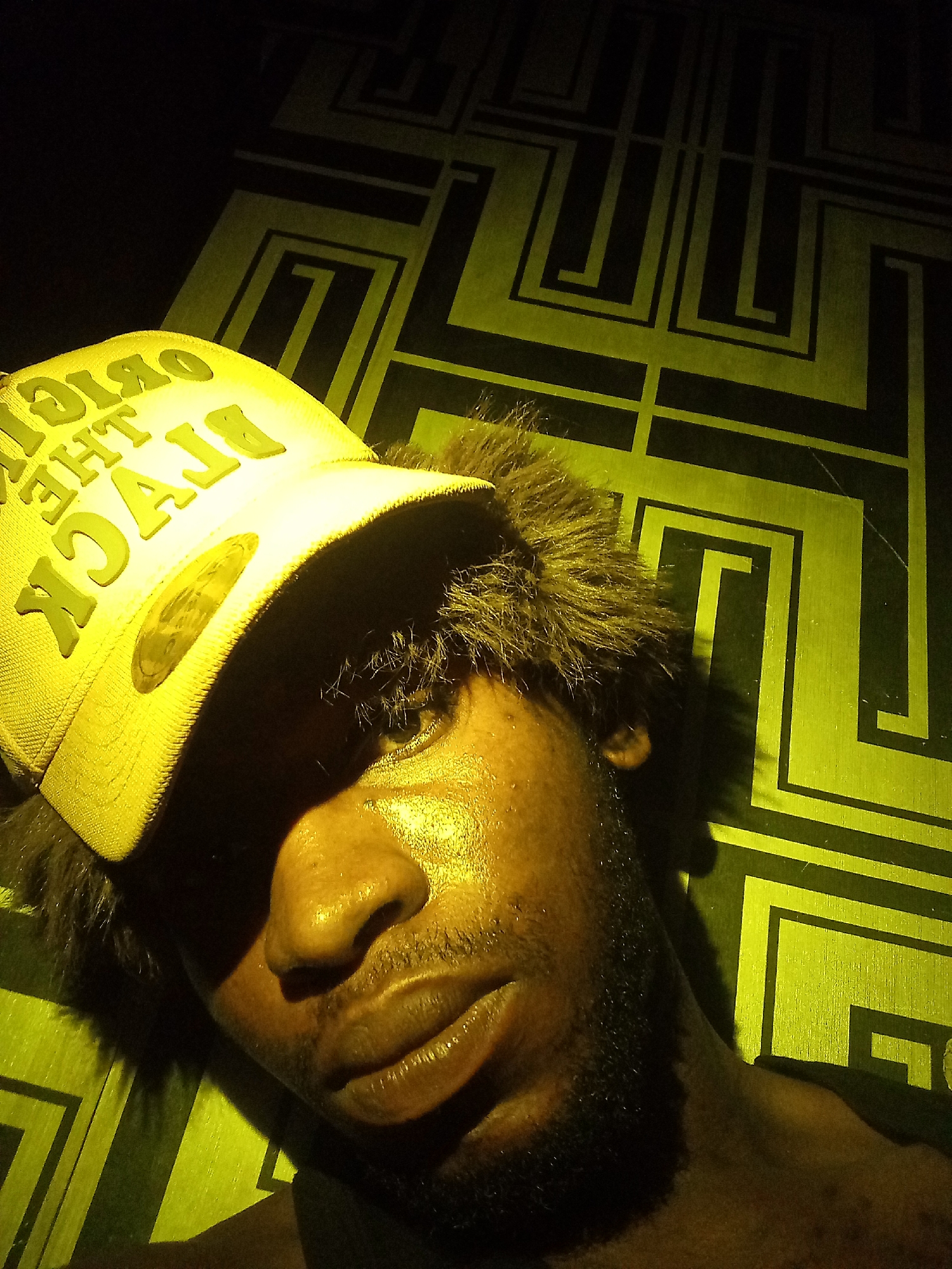 A close-up, high-angle selfie shows a man with dark skin wearing a yellow baseball cap and a beard. The cap has raised yellow lettering that reads "ORIGIN OF THE BLACK". His face is illuminated from above, casting shadows and highlighting the sheen of perspiration on his skin. The background consists of a dark surface with a repeating geometric pattern in a lighter shade of yellow, resembling a maze or labyrinth. The image appears to have been taken indoors, possibly in a room with low lighting and a unique wall decoration.