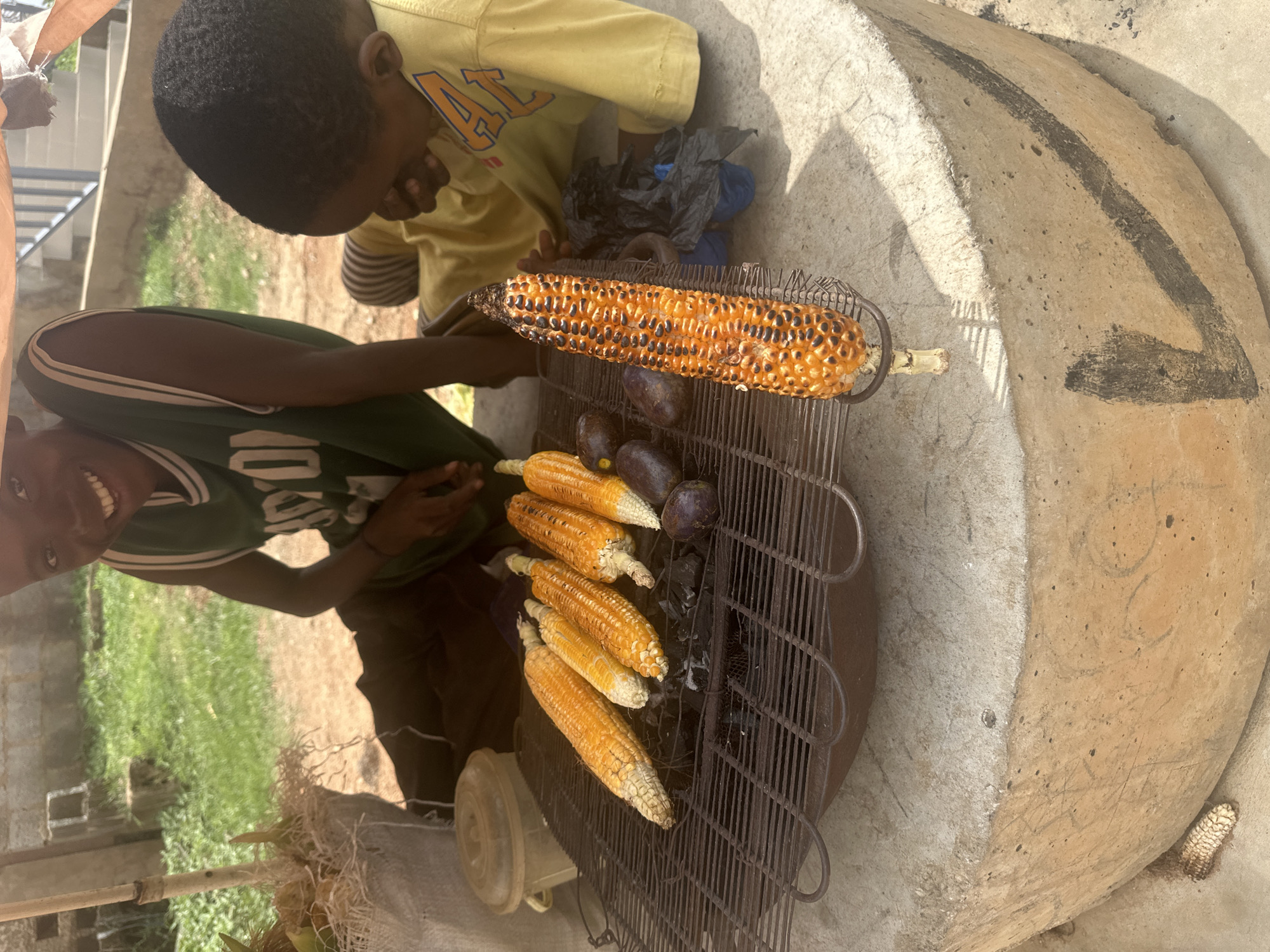 The image captures a street vendor scene in Gwarinpa, Nigeria, with two young boys present. In the foreground, a metal grill is placed on top of a large, round, concrete structure, likely an oven or brazier. The grill holds several ears of corn being roasted over coals, with one ear visibly charred. Adjacent to the corn are a few dark, oval-shaped items, possibly plantains or a similar produce. A white plastic container is visible to the left of the grill.
One boy, wearing a green basketball-style jersey, is seated behind the grill, smiling broadly at the camera. He appears to be actively involved in the roasting process, with his hands near the grill. Another boy, wearing a yellow t-shirt with orange lettering, is seated to the right, looking down and possibly interacting with the food. The background shows a dusty, outdoor environment with greenery and what appears to be a building with a metal railing. The overall impression is a candid street scene depicting the sale and preparation of roasted corn.