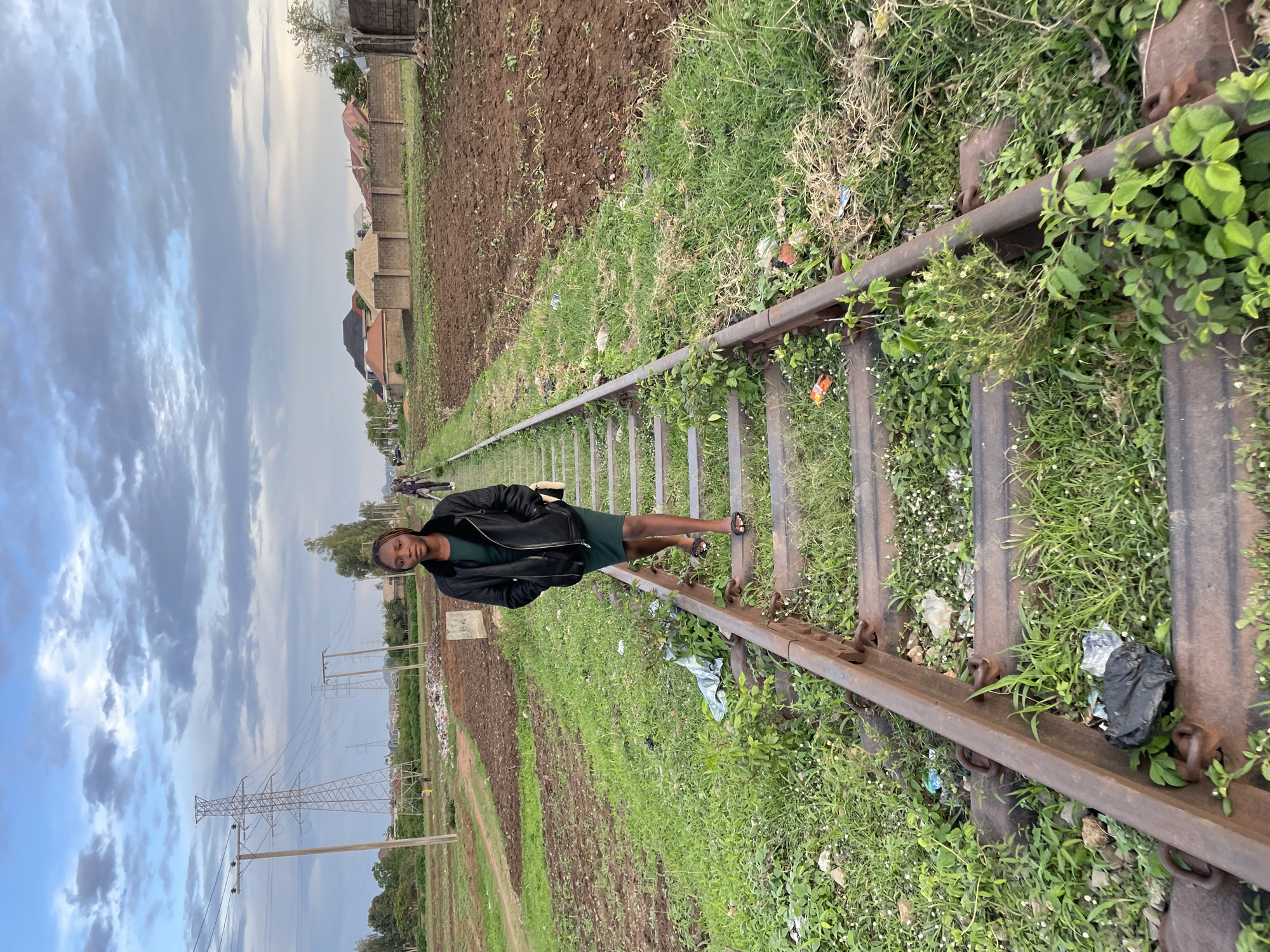 A young adult female walks towards the viewer on a single set of railway tracks in Jos, Nigeria. She has dark skin and wears a black jacket over a dark green dress, with black flat sandals and a light-colored head covering. Her hands are in her jacket pockets, and a small, light-colored bag hangs from her right shoulder. The railway tracks feature rusty rails and weathered sleepers, with green grass and various weeds growing between and around them. Scattered litter, including plastic bottles, wrappers, and a black plastic bag, is visible along the tracks. To the left of the tracks, cultivated fields with dark brown soil and green sprouts extend towards a series of wooden electricity poles and a large metal lattice transmission tower against a partly cloudy, light blue sky. To the right, similar cultivated fields are bordered by concrete and brick walls, behind which residential structures with varying roof colors are visible. Further down the tracks in the distance, another individual walks away from the camera.