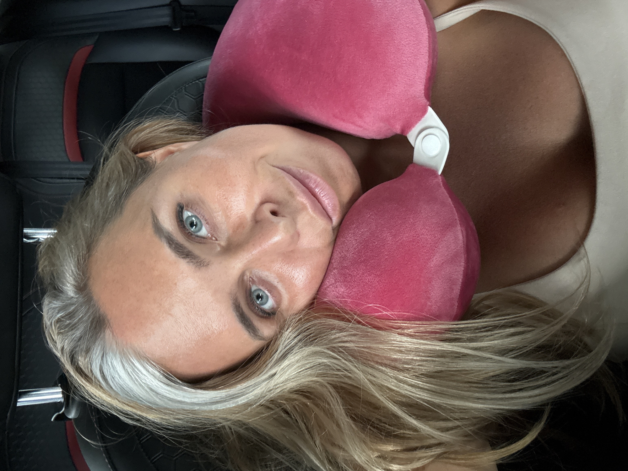 A woman with blonde and gray hair is resting her head on a pink neck pillow, looking at the camera. She is wearing a light-colored tank top and appears to be in a car. The background shows dark car seats with a red accent stripe.