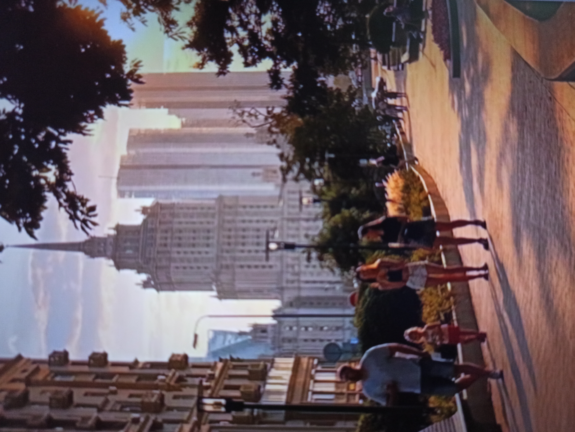 The image depicts a street scene in Balakliya, Ukraine, during what appears to be late afternoon or early evening, given the warm, low sunlight casting long shadows. A prominent Stalinist skyscraper with a spire dominates the background, flanked by modern glass skyscrapers. In the foreground, a paved walkway is lined with trees and planters. Several individuals are walking on the pathway. On the left, a man in a grey t-shirt and shorts is walking with a young child in a pink dress. To their right, two women are walking together; one has long blonde hair and is wearing a black crop top and white shorts, while the other has dark hair and is wearing a dark dress. Further down the walkway, other pedestrians can be seen in the distance. The architecture of the buildings and the overall atmosphere suggest a well-maintained urban environment.