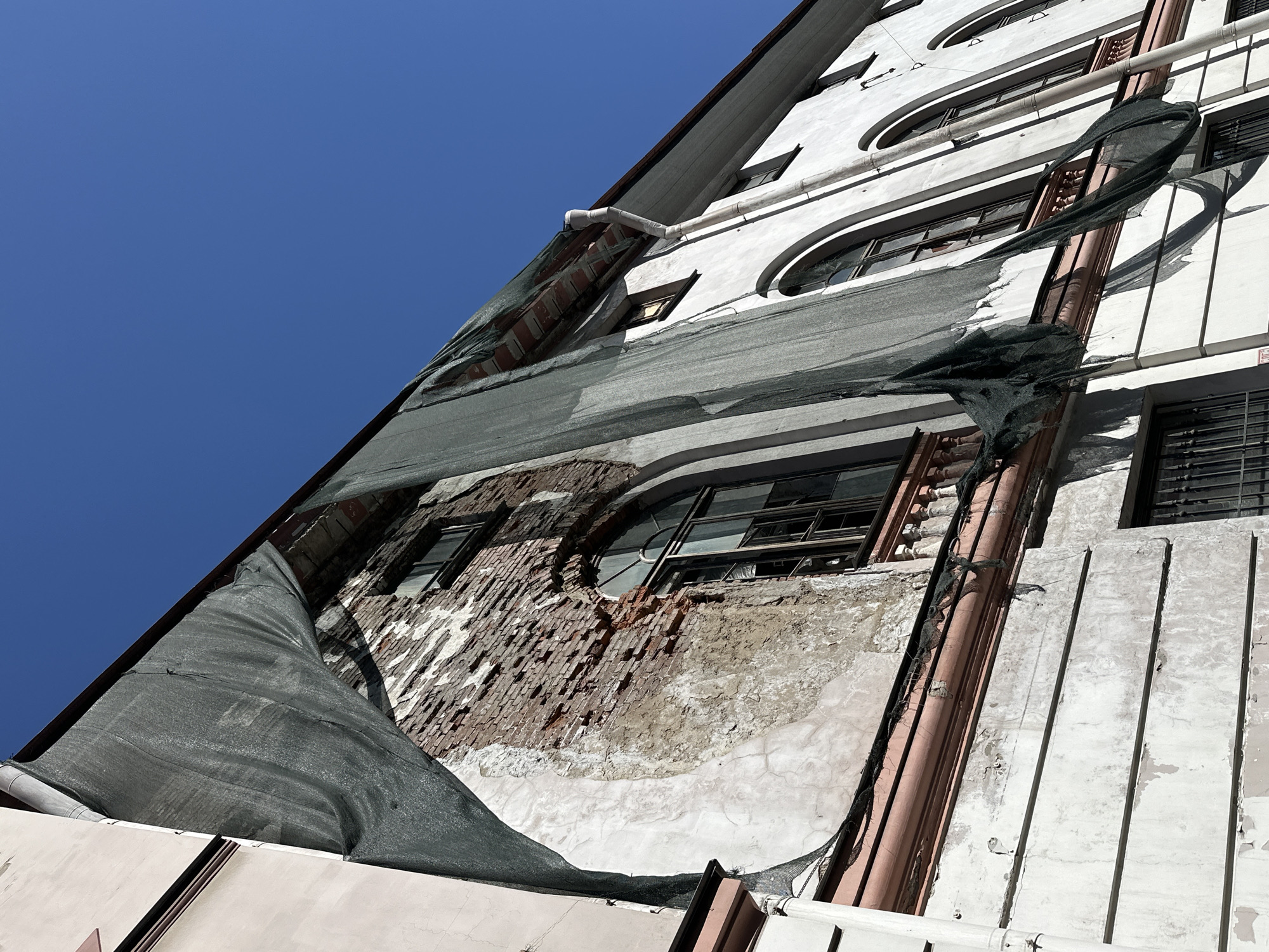 The image captures a dramatic, low-angle, and tilted perspective of a decaying building facade against a vibrant blue sky, suggesting a bright, clear day, likely midday due to the strong shadows. The architecture, with its mix of rectangular and arched windows, ornate moldings, and exposed brickwork, points to an older urban structure, consistent with the provided context of Saint Petersburg, Russia.
The building's exterior is predominantly covered in a light-colored render or plaster, much of which has peeled away, revealing the reddish-brown brick skeleton beneath. This significant deterioration is particularly noticeable in the central and lower-left areas of the frame. Several windows are visible, many appearing broken, missing panes, or covered with grates, emphasizing the building's neglected state.
A large section of dark green or black construction safety netting is draped across the facade. It appears torn and billows outward in places, casting dynamic shadows and partially obscuring the underlying brickwork and windows. This netting suggests either an ongoing renovation project, a stalled repair effort, or a safety measure to contain falling debris from the crumbling edifice.
Various utility pipes, some white and others a reddish-brown, run both vertically and horizontally along the building's exterior, adding to the intricate textures of the scene. The clear, deep blue sky fills the upper-left portion of the frame, providing a stark contrast to the weathered and dilapidated structure. There are no people or discernible text visible in the image. The overall impression is one of urban decay, the passage of time, and the remnants of past architectural grandeur.