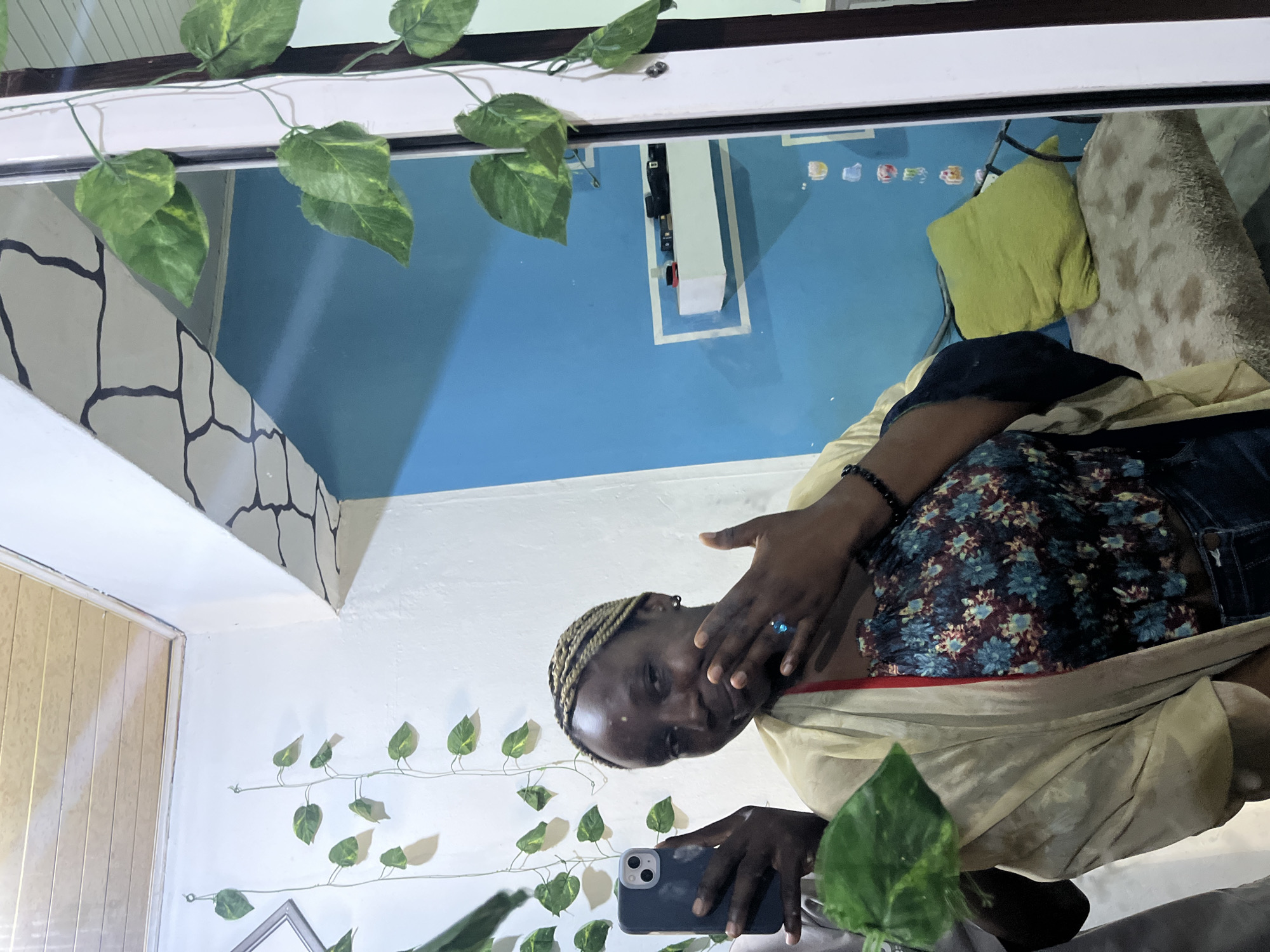 A person is taking a selfie using a smartphone in front of a mirror. The mirror reflects a room with blue walls, a white fixture, a green pillow, and a beige cushion. Green vines are draped along the top and left side of the mirror. The person is wearing a floral top, a light-colored shawl, and a black bracelet. They are holding the phone in their left hand and touching their face with their right hand. The location is Unknown, Unknown.