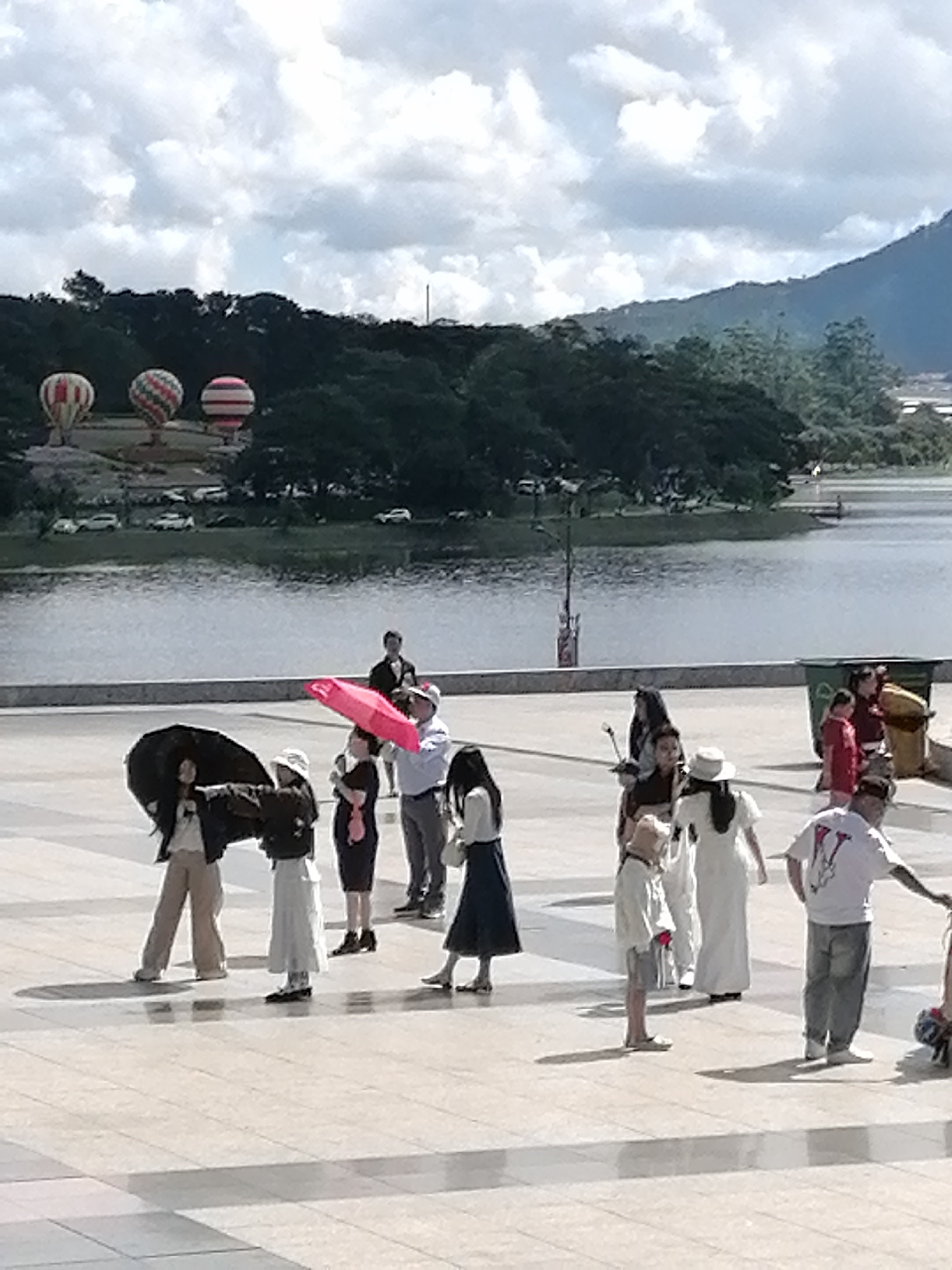 The image captures a daytime scene at Xuan Huong Lake in Da Lat, Vietnam, with a group of people gathered on a paved plaza. In the background, three hot air balloons are visible against a cloudy sky, positioned above a tree line that borders the lake. The lake itself is calm, reflecting the sky and trees.
In the foreground, several individuals are interacting. One person, wearing a black jacket and tan pants, is holding a black umbrella open. Another person, in a white skirt and dark top, stands beside them. A third person, wearing a white shirt and dark pants, is holding a bright pink umbrella aloft, seemingly pointing. Other figures in the background include individuals in white dresses, a person in a white t-shirt and jeans, and a child in a light-colored outfit and shorts. Some individuals appear to be tourists, possibly preparing for or just having participated in an activity related to the hot air balloons. The plaza is paved with square tiles, and shadows are cast by the people, indicating sunlight. To the right, a green trash can is visible. The overall atmosphere suggests a leisurely outdoor activity or event in a picturesque setting.