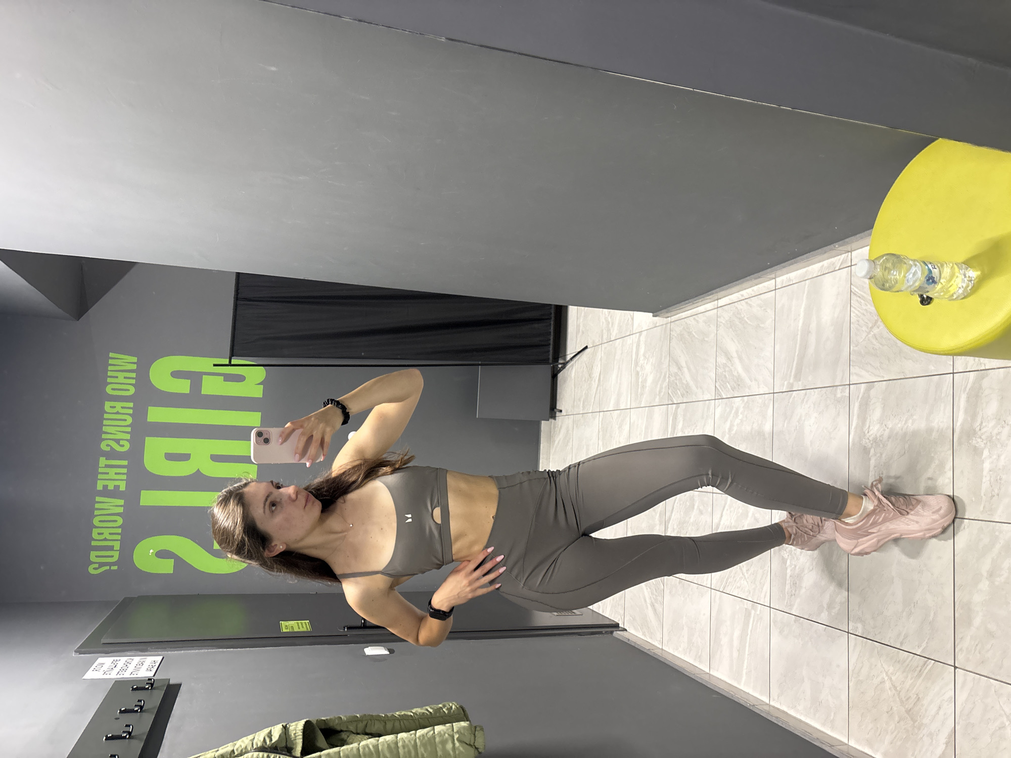 A woman is taking a selfie in a gym changing room. She is wearing a grey sports bra and matching leggings, and pink sneakers. She has her hair pulled back in a ponytail and is wearing a small necklace. In the background, there is a wall with a green motivational quote and a black curtain. There are also hangers on the wall and a green jacket hanging on one of them. A yellow stool with a bottle of water on it is visible in the foreground. The floor is tiled. The image was taken in the city of Varna, Bulgaria.