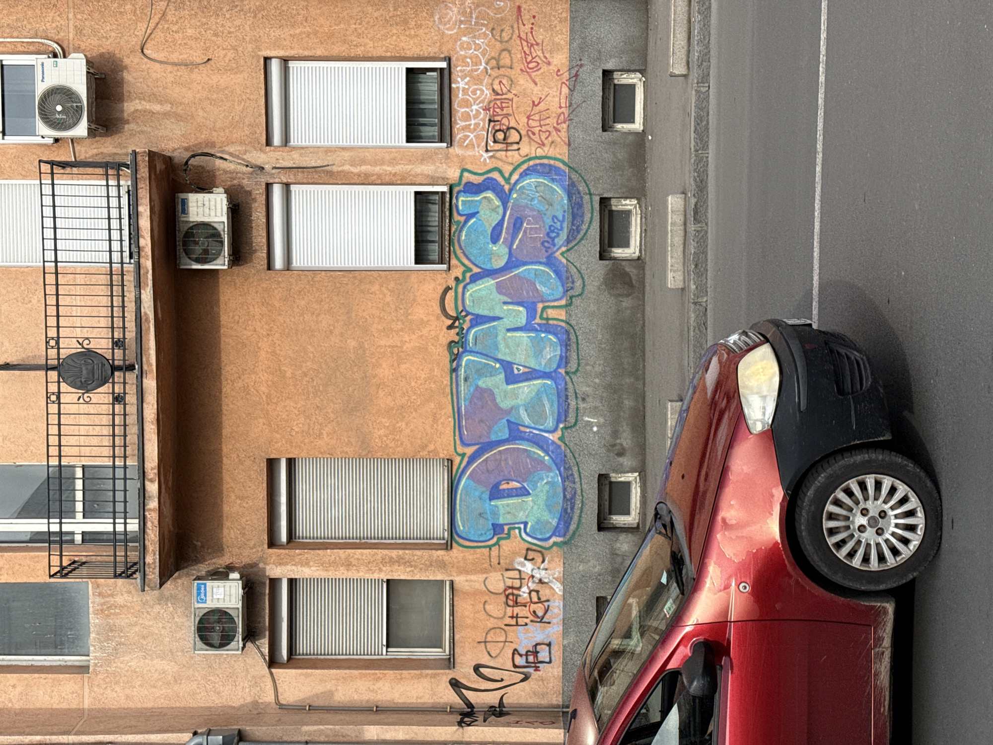 An eye-level shot shows the side of a building and a parked car. The building's facade is divided vertically into two sections, one section beige and the other gray. Graffiti art is spray-painted on both building sections. Each section has four evenly spaced window openings and a window unit in each opening. There are four air conditioning units affixed to the beige section of the facade. A black fire escape is attached to the beige building section. A red car with a black hood is parked on the gray street in front of the building. The location is Vracar Urban Municipality, Serbia.