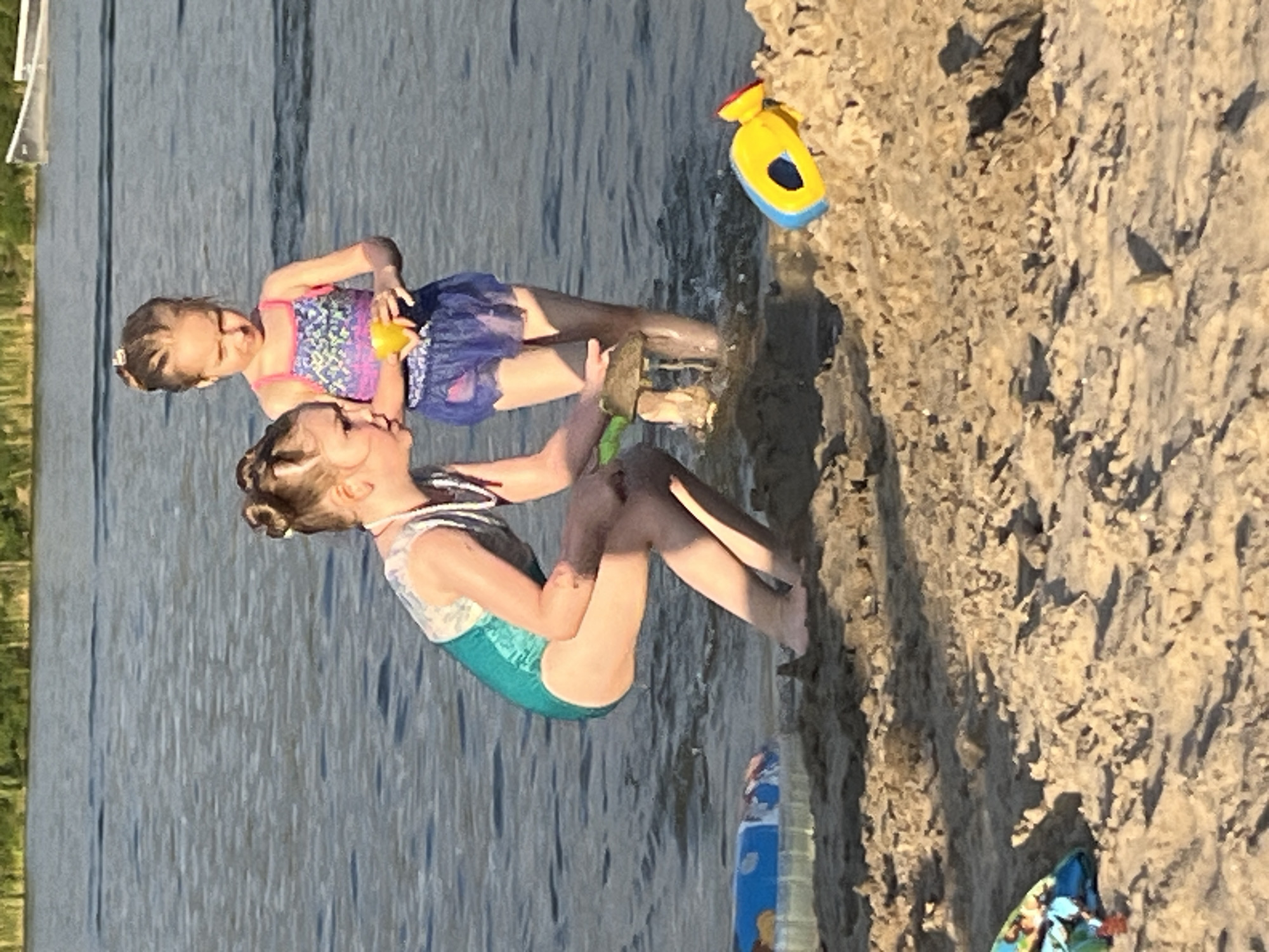 Two young girls are playing on the shore of a body of water, likely a lake or river. One girl in the foreground, wearing a teal and white swimsuit, is sitting on the sandy bank, holding a small toy. She has her hair pulled up in a bun and is looking down, perhaps at the toy. The second girl, in the background, is wearing a pink and purple swimsuit with a blue tutu. She is also in the water, holding a yellow toy. Her hair is also in a bun. The scene appears to be set in the late afternoon or early evening, given the long shadows cast on the sand. The water is calm, reflecting the sunlight, and the sand is a light brown. In the background, a blurry green area suggests vegetation. There is a yellow and blue toy floating in the water. The overall mood is playful and relaxed, suggesting a summer day spent at the beach. There is no visible text in the image. The location is specified as Dnipro, Ukraine.