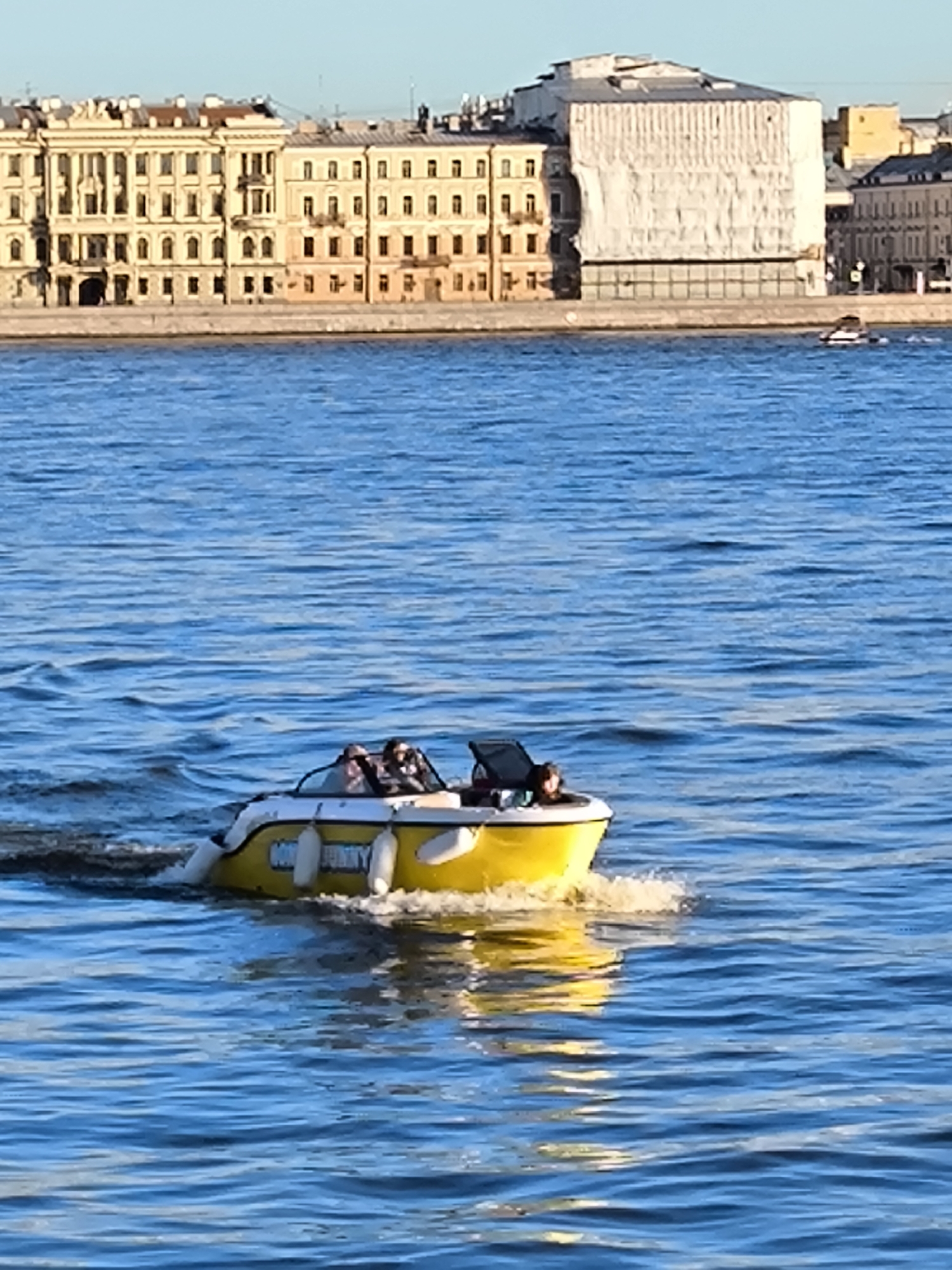 A bright yellow motorboat with a white top is traveling on a wide, blue body of water, likely a river or canal in Saint Petersburg, Russia. Three people are visible in the boat, seated and facing forward, appearing to be enjoying a ride. The boat is leaving a wake behind it. In the background, across the water, is a long row of historic buildings with light-colored facades and multiple windows, characteristic of Saint Petersburg's architecture. Some of these buildings are adorned with classical architectural elements. One large building in the mid-background is covered in white sheeting, suggesting it may be under renovation or maintenance. The sky above is clear and a light blue.