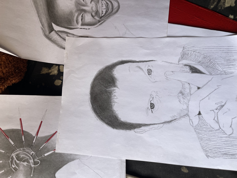 This image displays a collection of three pencil drawings on white paper sheets, arranged on a dark, textured surface, likely in a studio or workspace in Jos, Nigeria.
The most prominent drawing, centrally located, is a detailed portrait of a light-skinned male. He has short, dark hair, stubble around his jawline, and is depicted with his left index finger pressed vertically against his lips, indicating a "shhh" gesture. His eyes are narrowed, directed forward, and he is wearing a vertically striped garment.
To the top left, a partial view of another drawing is visible. This artwork features a human ear, meticulously rendered, adorned with multiple sharp, pointed objects that have distinct red tips, suggesting a form of body art or piercing. The drawing is predominantly monochromatic with selective red coloration.
In the upper right quadrant, another partial drawing shows a smiling person, possibly a child or young adult, with dark skin. The individual's wide smile reveals teeth, and a portion of a hand is visible near their head.
The drawings are laid on a dark, irregular surface that shows some lighter specks and shadows, with hints of red and other dark patterns in the periphery, suggesting a varied background material.