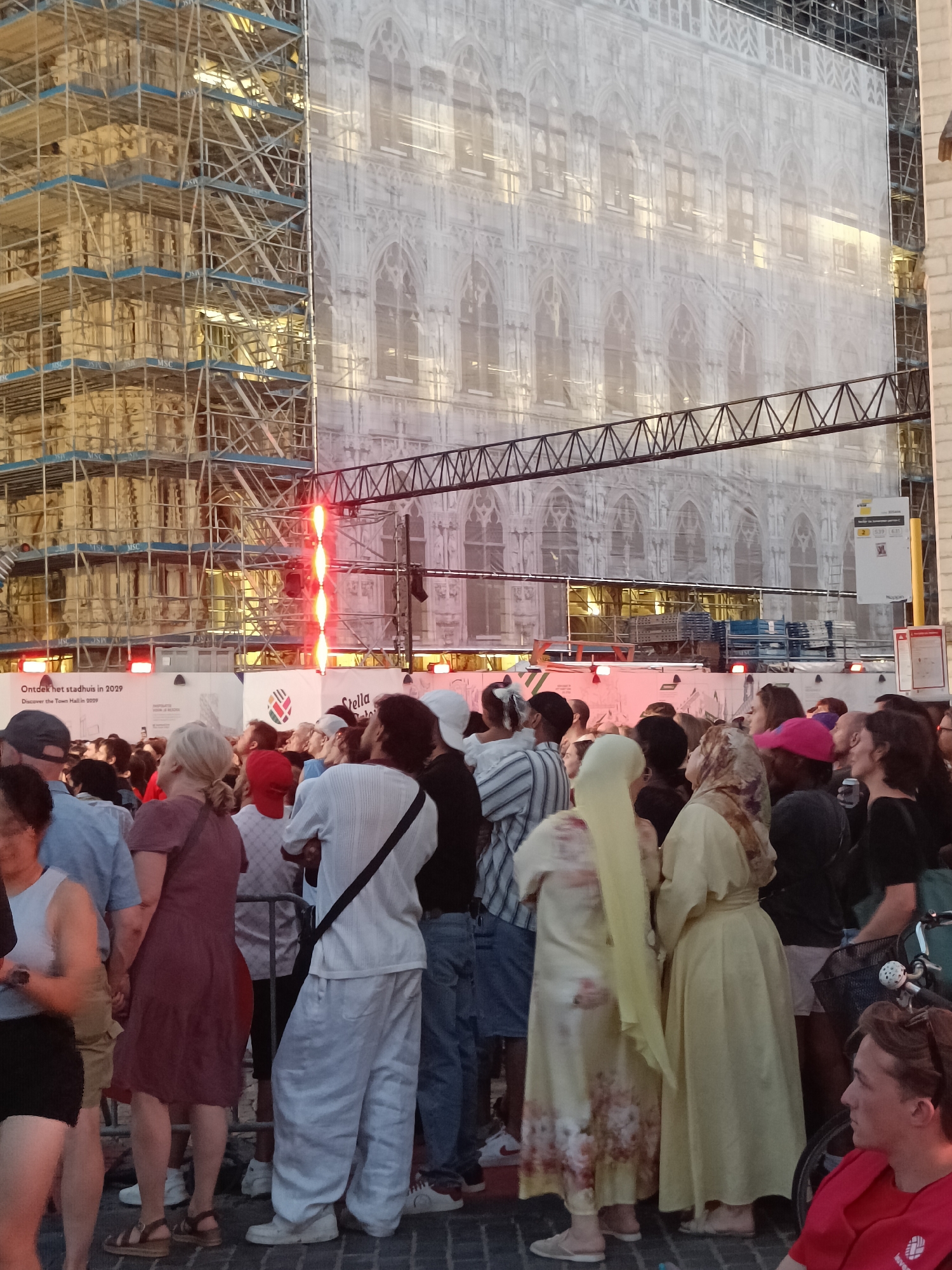 The image depicts a crowd of people gathered in front of a building covered in scaffolding and a large white tarp, suggesting ongoing construction or renovation. The tarp displays a printed image of the building's architectural details, creating an illusion of its original appearance. Red lights are prominently displayed on scaffolding, potentially for illumination or safety. A banner at the bottom of the frame reads "Ontdek het stadhuis in 2029 Discover the Town Hall in 2029", indicating the location is Leuven, Belgium, and the building is the town hall. People are dressed in casual attire, with some looking towards the building, while others are engaged in conversation or looking elsewhere. The ground is paved with cobblestones. In the background, more scaffolding and construction materials are visible.