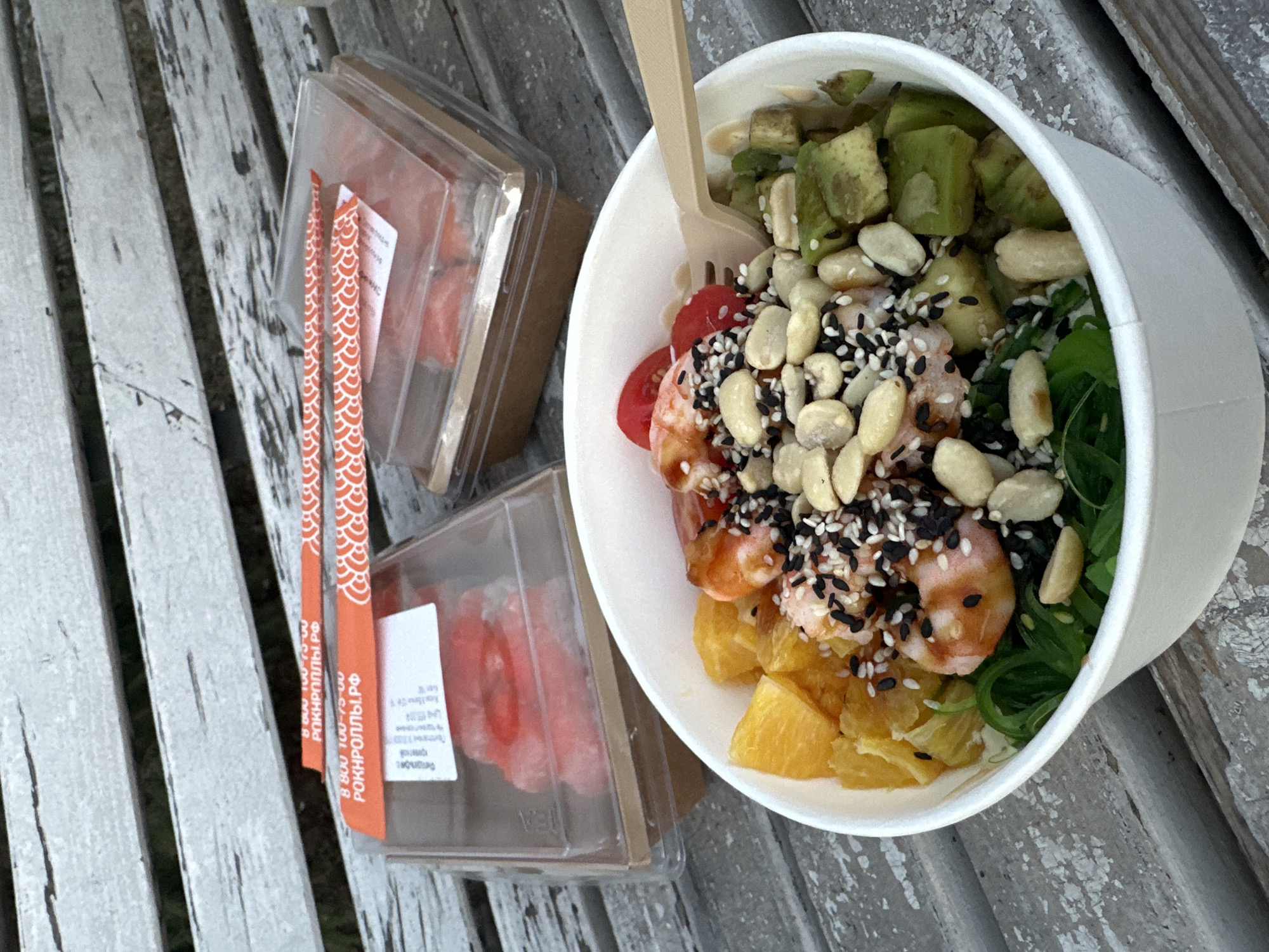 The image shows a close-up of food on a weathered wooden surface, likely a bench. Two clear plastic containers, potentially holding sushi rolls, are visible on the left side. Next to them is a white bowl filled with a variety of ingredients including diced avocado, tomatoes, shrimp, diced pineapple, seaweed and peanuts topped with black sesame seeds. A light brown disposable fork rests inside the bowl. Chopsticks with a red and white pattern are also visible between the sushi containers. The text "8 800 100-75-00 РОКНРОЛЛЫ.РФ" is printed on the chopstick wrapper. The setting suggests an outdoor location. It is not possible to verify if this image was taken in Kazan, Russia from the provided information.