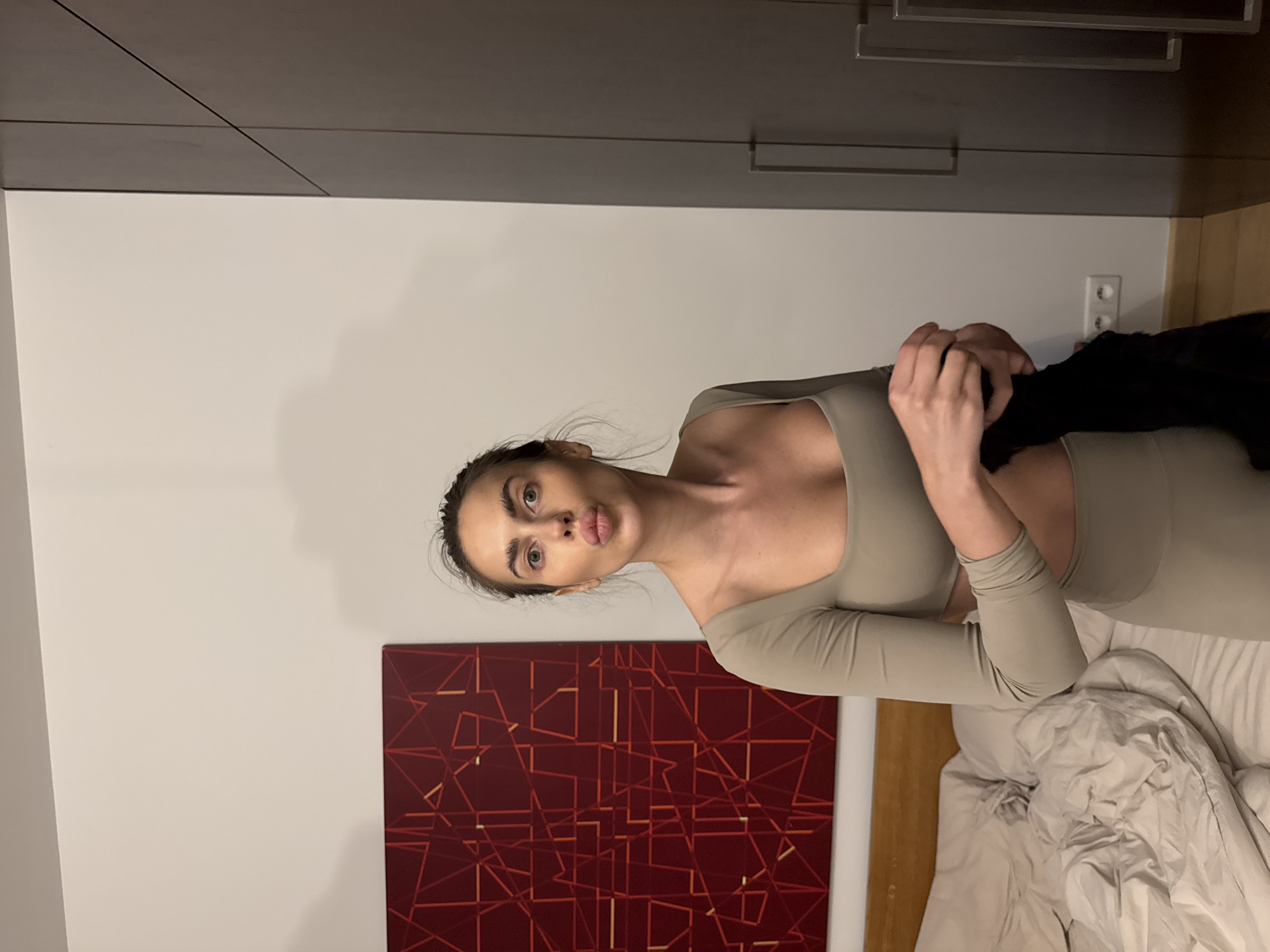 A woman is holding a black item of clothing while sitting on a bed. She is wearing a light-colored, form-fitting outfit. Behind her is a white wall with a red abstract painting. Further back, there are light-gray cabinets and a wall outlet. The location is likely indoors. The city of Warsaw, Poland, cannot be confirmed.