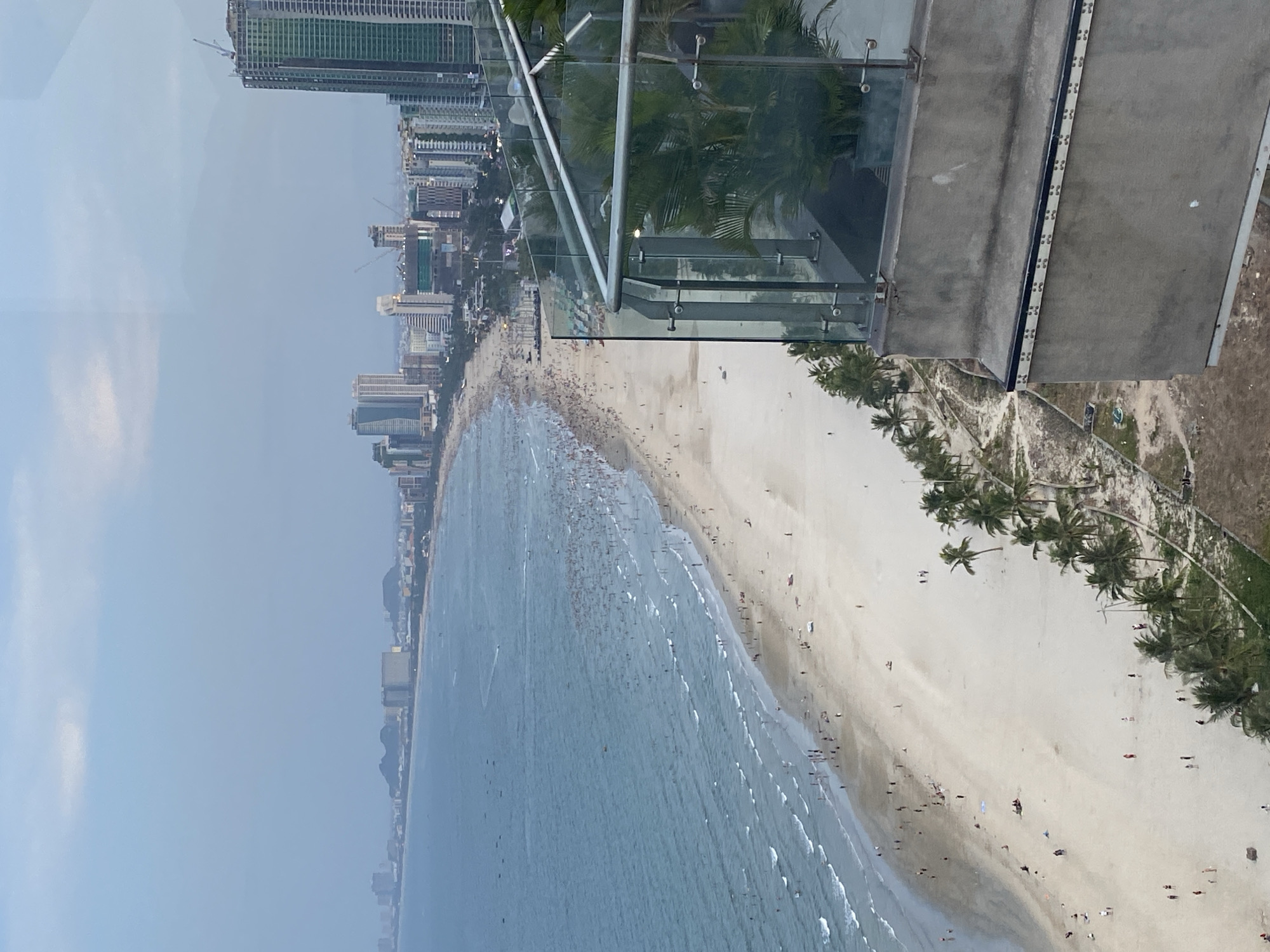 This is an aerial view of My Khe Beach in Da Nang, Vietnam, likely taken from a high-rise building. The scene is dominated by a vast expanse of sandy beach and the turquoise waters of the sea. A large number of people are visible on the beach, spread out across the sand and wading in the shallow waters, suggesting a busy day. The water is dotted with small boats and a few individuals engaged in water activities.
In the background, a densely populated urban landscape stretches along the coastline, characterized by numerous high-rise buildings, including a prominent green-tinted skyscraper under construction. The city skyline suggests a thriving coastal city with modern development. Palm trees line the edge of the beach, adding a tropical element to the scenery. In the foreground, a concrete balcony with a glass railing, furnished with potted palm plants, frames the view. The sky is overcast with a hazy, soft light. The overall impression is one of a popular tourist destination with a lively atmosphere.