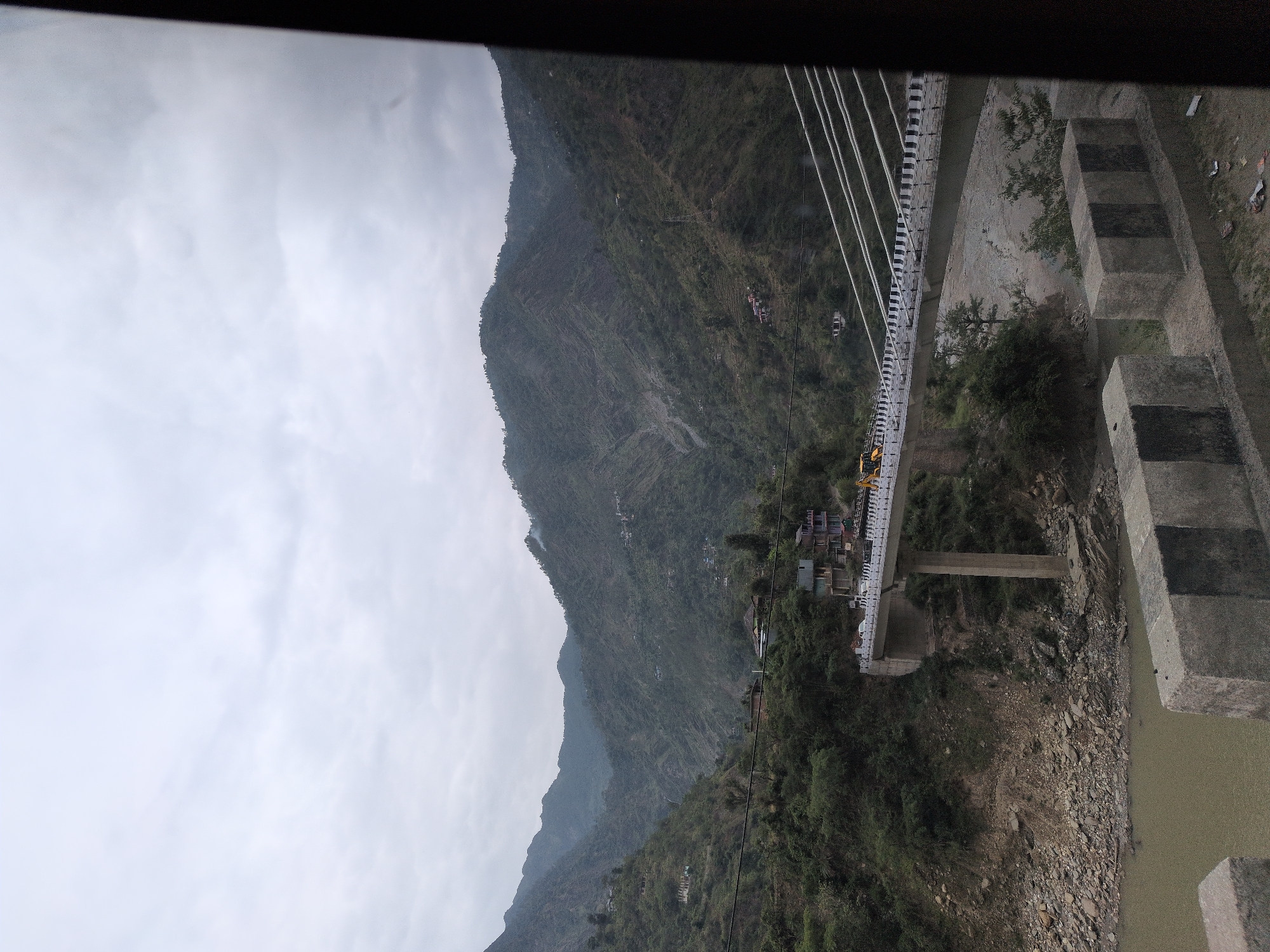 The image captures a suspension bridge spanning across a river valley in Hanogi, India. The bridge, a significant piece of infrastructure, features multiple white cables supporting its deck, which is lined with black and white reflective markers and a railing. Several vehicles, including cars and a yellow construction vehicle, are visible on the bridge. The surrounding landscape is mountainous and heavily vegetated, with dense green trees covering the slopes. Small settlements with houses are scattered across the hillsides. The river below the bridge appears muddy and flows through a rocky terrain. The sky is overcast with grey clouds, suggesting a cloudy or possibly rainy day. The foreground shows concrete barriers, likely part of a road or walkway. The overall scene depicts a rural mountainous environment with human development in the form of the bridge and scattered settlements.