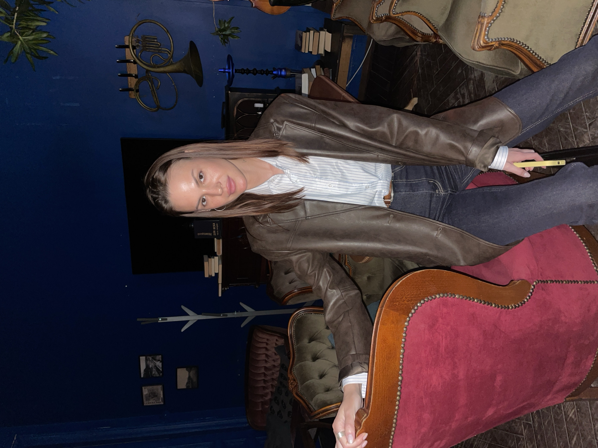 A woman sits in a richly decorated interior space, likely in Saint Petersburg, Russia, indicated by the classic, ornate furnishings and decor elements. She is wearing a brown leather jacket over a blue and white striped shirt, and dark jeans. In her right hand, she holds a yellow smartphone. The surrounding furnishings include velvet chairs with decorative nailhead trim, a hookah, and wall decorations like a French horn. The walls are painted a deep blue.