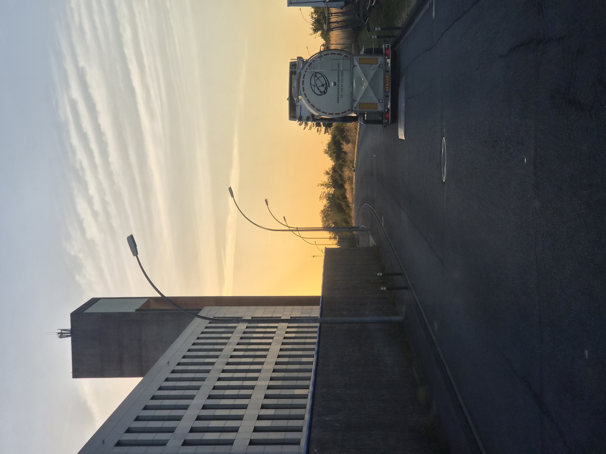 The image depicts a road in Leudelange, Luxembourg, during what appears to be sunset or sunrise, with a warm orange glow on the horizon. On the left side of the road, there is a modern, grey building with a repetitive pattern of windows and a tall, concrete tower with antennae on top. A dark, textured wall runs along the side of the building. The road itself is asphalt, with visible cracks and a manhole cover.
On the right side of the road, a white tanker truck is parked. The truck has a circular logo and text that reads "www.interkull-spedition.com Tel. +49 40 6391880". The license plate is visible and reads "LA 5013". Beyond the truck, there are trees and vegetation. Several streetlights with curved arms are positioned along the road. The sky is a pale blue with wispy, horizontal clouds. The scene suggests an industrial or commercial area on the outskirts of the city.