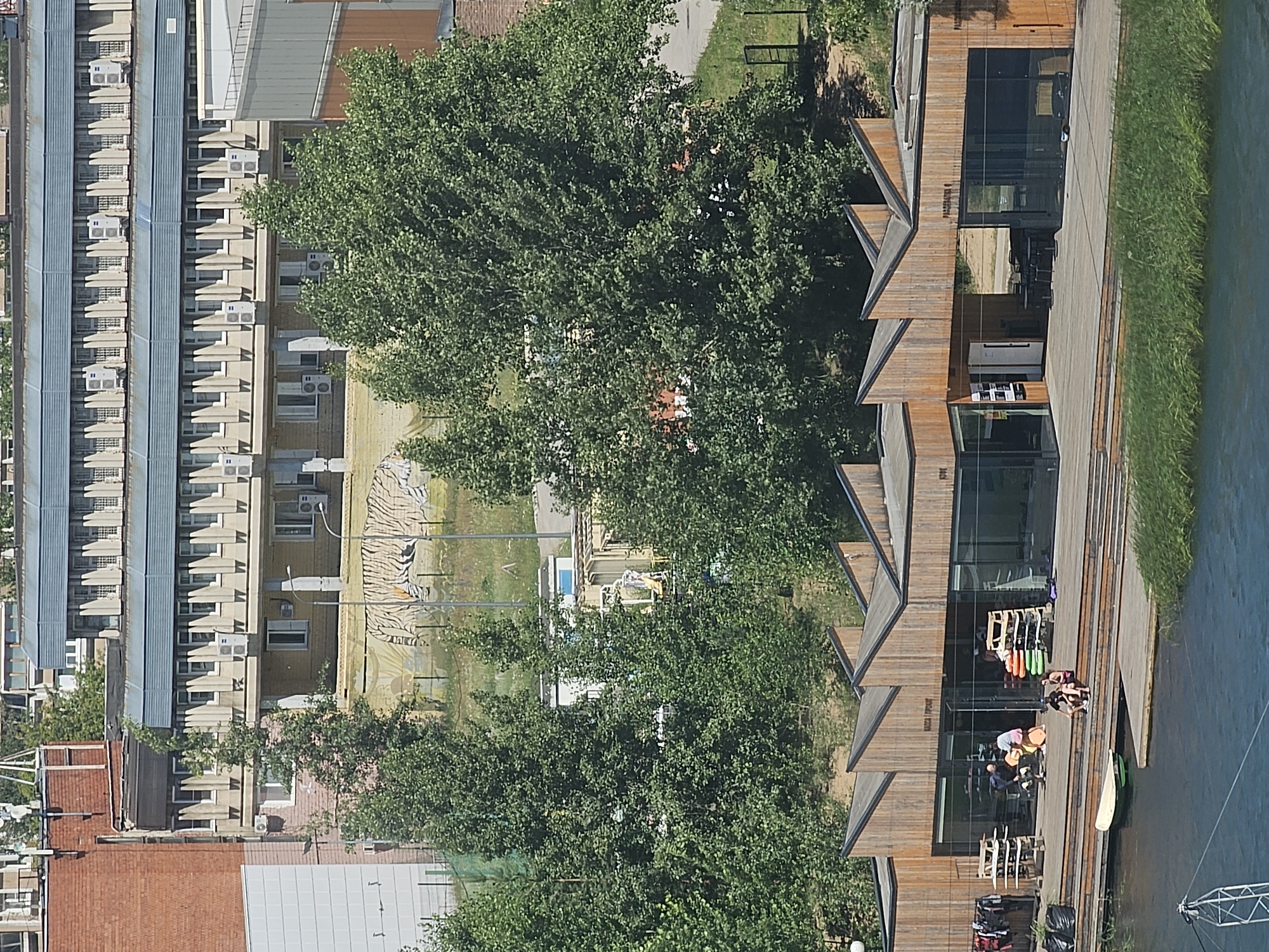 This is an aerial, slightly tilted, wide-angle photograph of an outdoor recreational area by a body of water. The scene is captured on a bright, sunny day. The primary focus is a modern, wooden building with extensive glass windows, situated on a deck overlooking the water. The building appears to be a cafe or club, with several tables and chairs visible both inside and on the deck. Near the water's edge, there are a few individuals engaged in activities, possibly preparing for or returning from water sports. Equipment like kayaks and what appears to be wakeboarding gear is visible.
To the left of the wooden building, a lush green tree provides shade. Beyond the tree, a series of large, multi-story buildings with repeating architectural features, possibly apartment complexes or office blocks, line a street. These buildings have many windows and air conditioning units. Further in the background, a large, unfinished-looking area with some machinery and a striped mural (resembling a tiger's stripes) is visible.
A prominent piece of text is visible on the wooden building, above the entrance: "КАЗАЧЬЯ ПРИСТАНЬ" (KAZACH'YA PRISTAN'). The presence of a body of water and the recreational nature of the scene suggests this is a popular spot for leisure activities. The bright sunlight indicates it is likely daytime, possibly late morning or afternoon. The overall mood is one of outdoor recreation and relaxation.