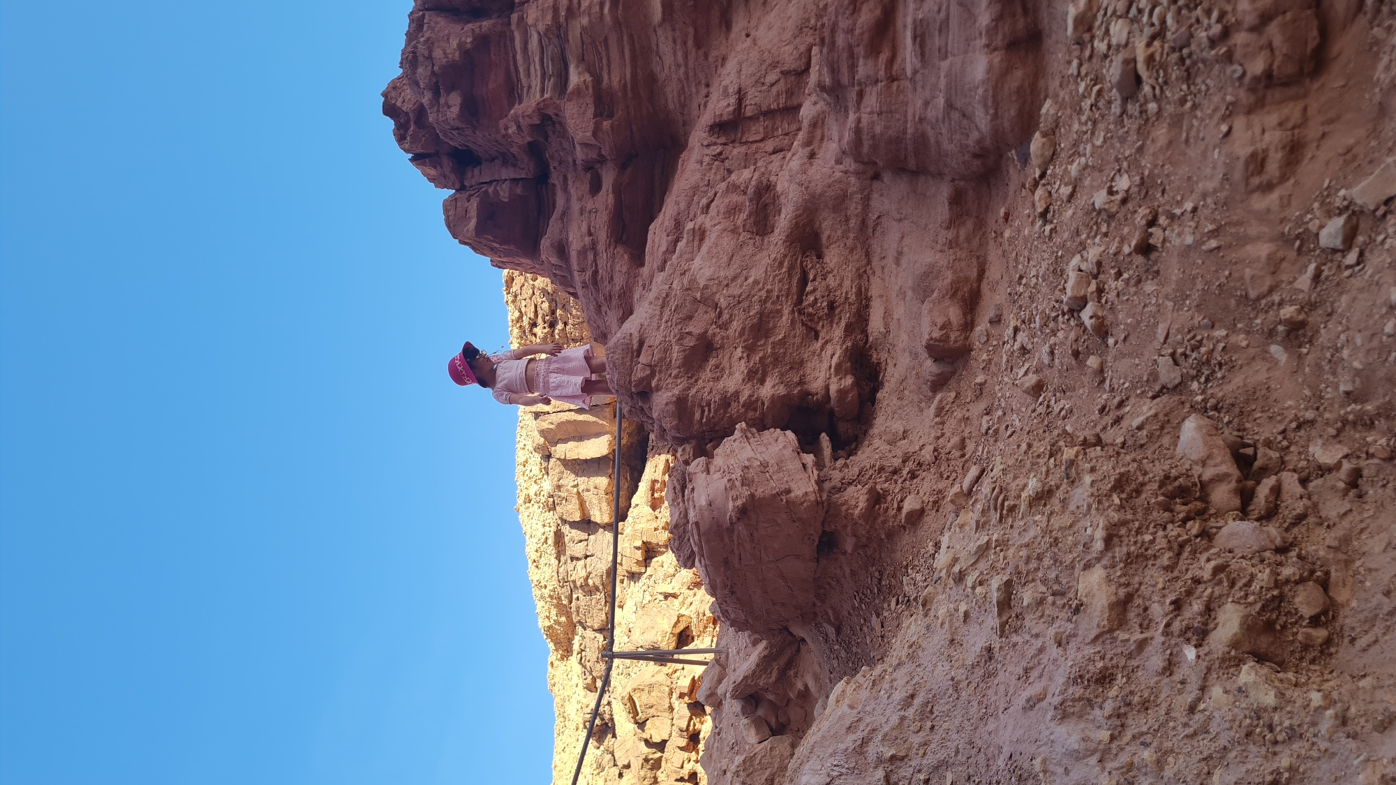 A person stands on a ledge halfway up a reddish-brown rock face against a clear blue sky. The person is wearing light-colored clothing and a red cap. The rock face appears to be part of a cliff or canyon wall, with varying textures and geological formations. A metal railing or support structure is visible along the ledge. The location is in Hevel Eilot Regional Council, Israel.