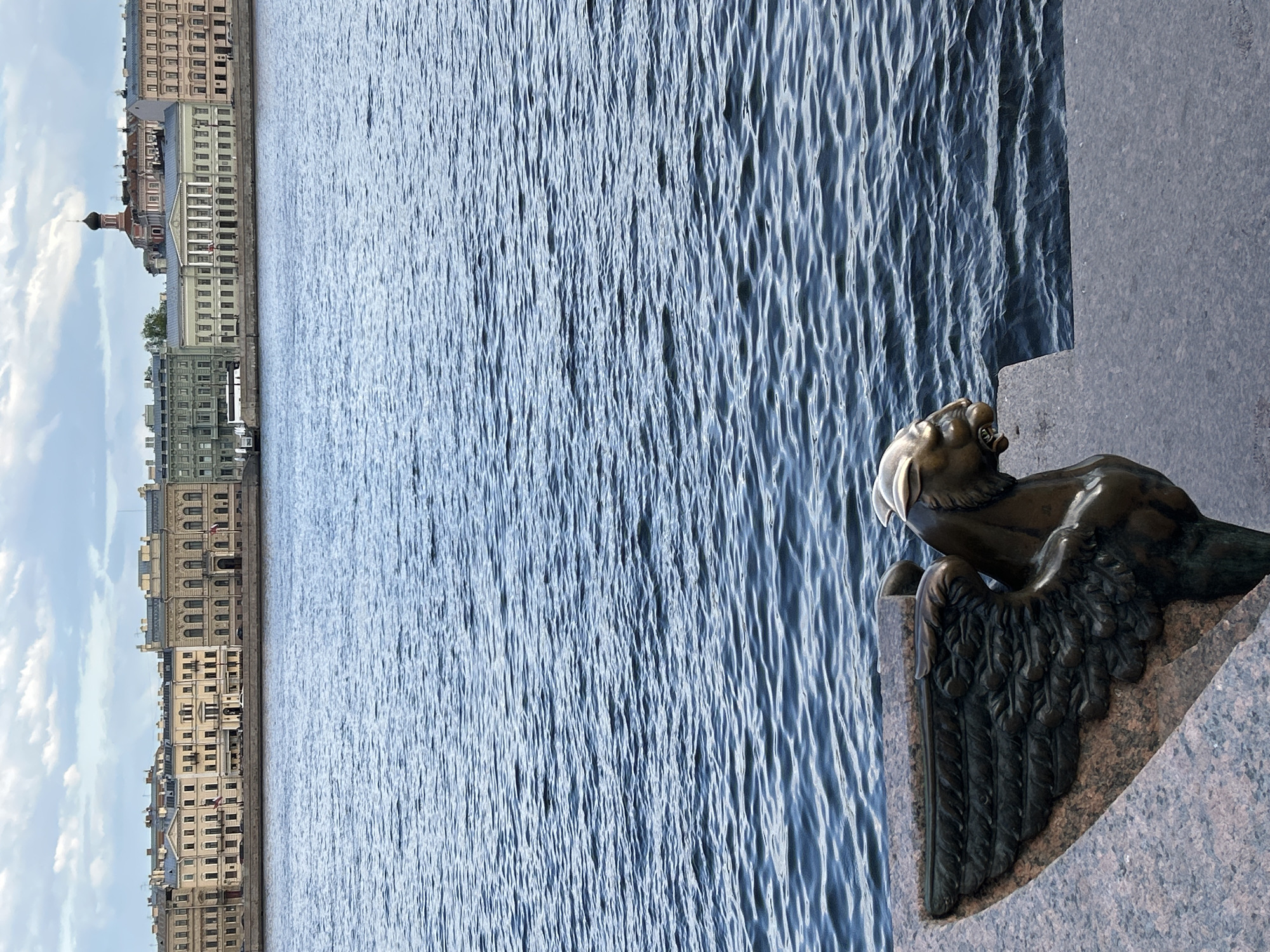 The image shows a sphinx statue on the embankment of the Neva River in St. Petersburg, Russia. The river occupies the center of the frame with the buildings and architecture of the city on the left side. A sphinx sits on the right. The image has been rotated 90 degrees counterclockwise.