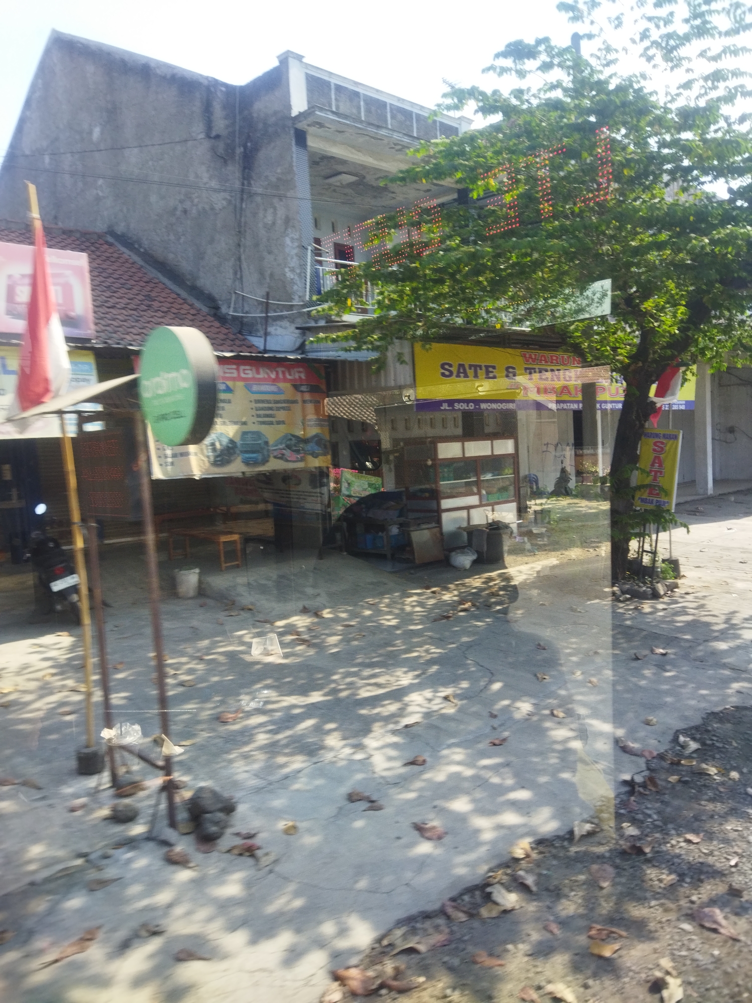 The image shows a street scene in Nguter, Indonesia. A building with a gray facade and a tiled roof is visible, with a second story that appears to be an outdoor balcony or walkway. A large tree with green leaves dominates the right side of the frame, its branches partially obscuring a string of red lights. Below the tree, a yellow sign reads "Sate & Tongseng" in Indonesian. To the left of the building, another yellow sign is visible with text that includes "S GUNTUR". A red and white flag is attached to a pole in front of the building.
In the foreground, a paved area with scattered leaves is visible. A motorcycle is parked on the left side, and there are various objects on the ground, including what appears to be a trash can and some discarded plastic bottles. The scene is well-lit, suggesting it is daytime. The overall impression is of a commercial establishment on a street in a populated area.