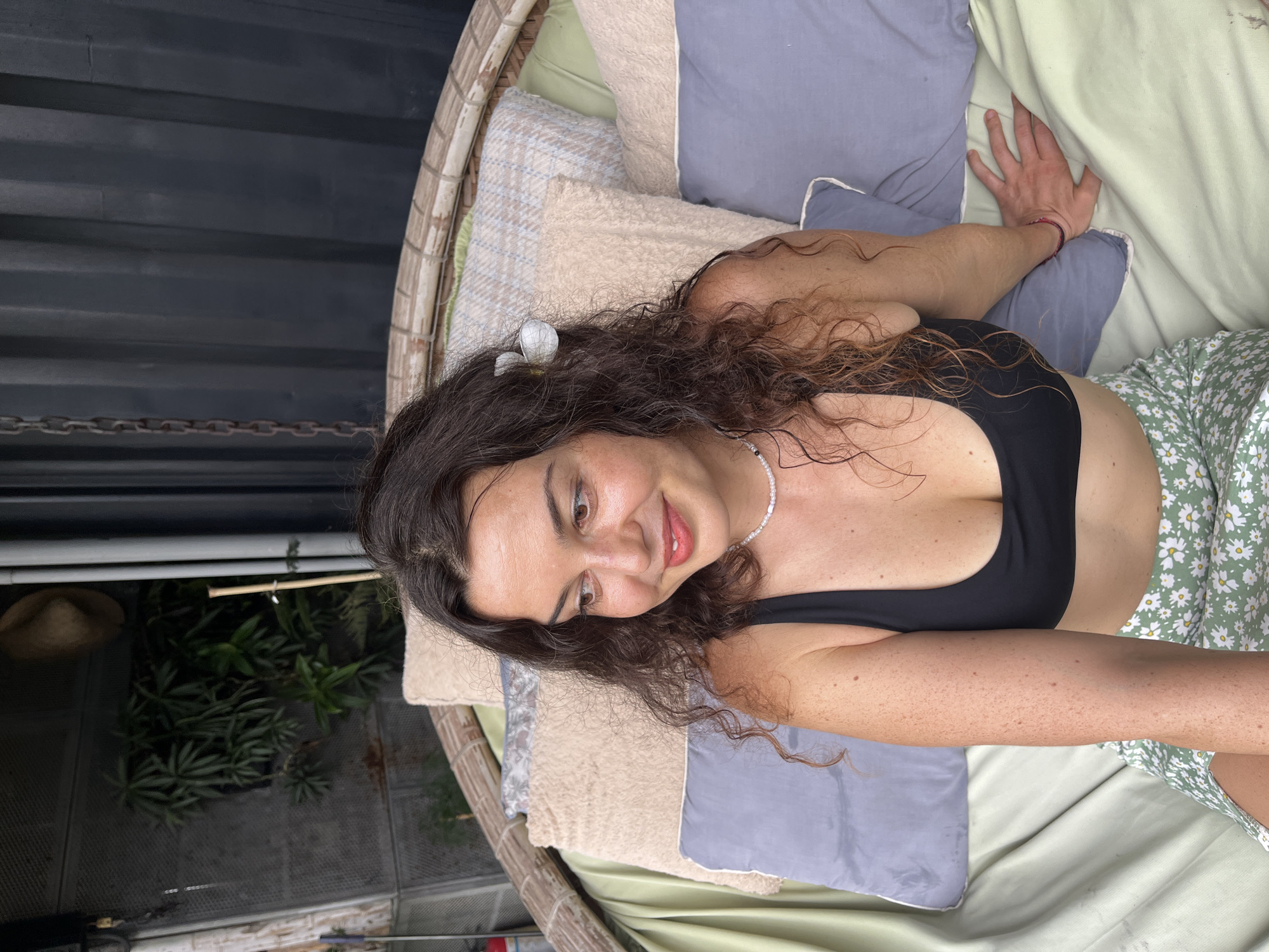 A woman with long, curly brown hair and a flower tucked behind her ear sits on a cushioned swinging chair outdoors. She is wearing a black bikini top and a green skirt with a white daisy pattern. She is looking to her left with a slight smile. The swinging chair is suspended from a dark, corrugated metal structure, and behind it, there are plants and a straw hat hanging on a wall. The scene appears to be in a relaxed, outdoor setting, possibly a patio or garden.