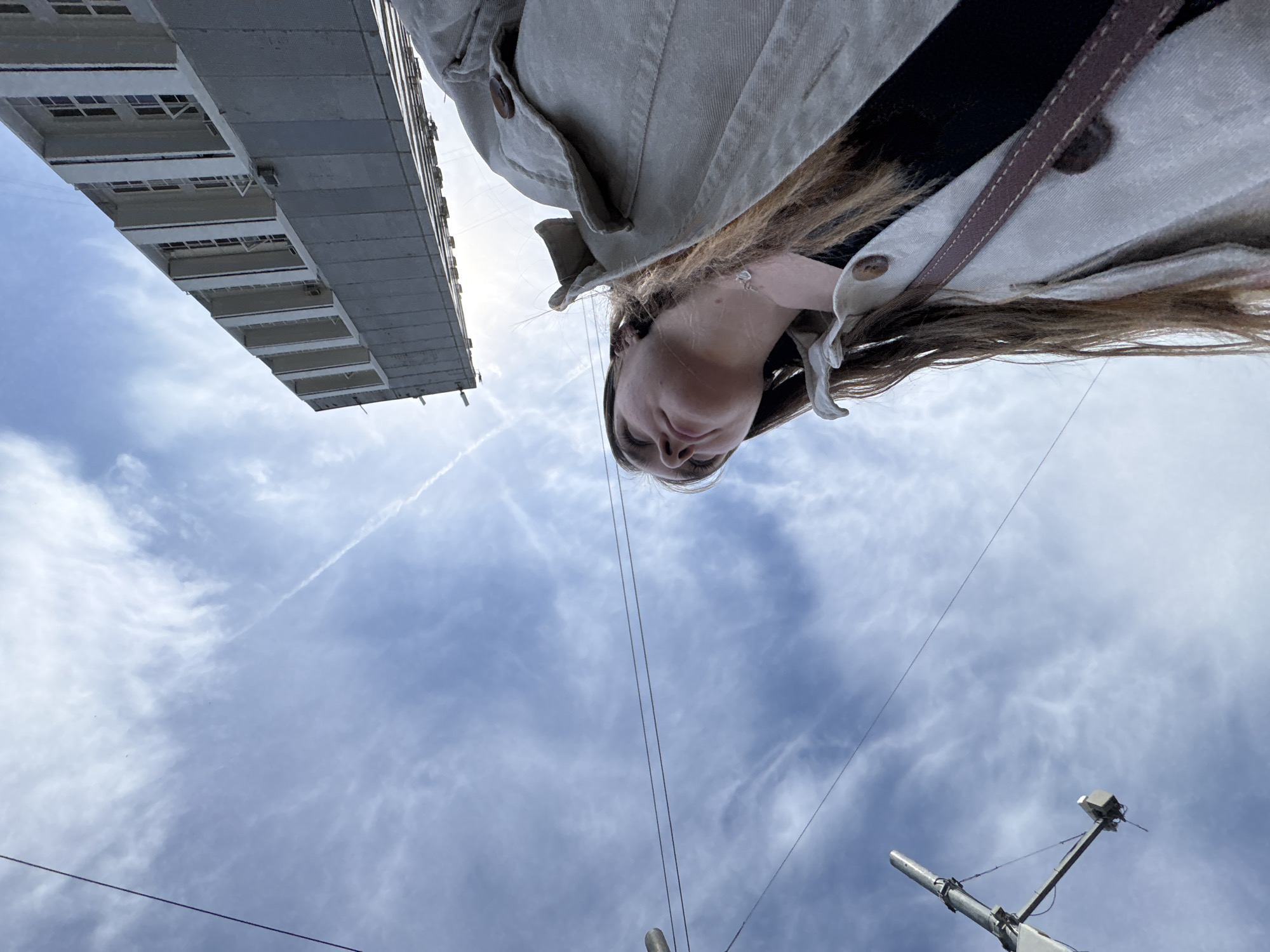 A low-angle shot shows a woman looking down at the camera while standing on a street. She has long brown hair and wears a light-colored jacket. In the background is a cloudy sky, part of a building, and an antenna. The location is Perm, Russia.