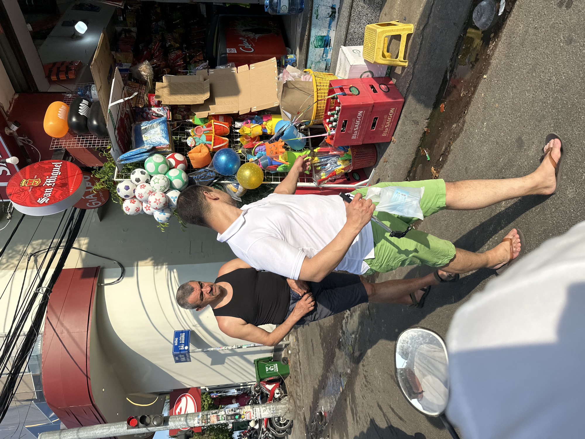 The image depicts a street scene in Bac Nha Trang Ward, Vietnam, showcasing a roadside stall laden with merchandise. Two men are present; one in a white polo shirt and green shorts appears to be interacting with the stall's offerings, while another man in a black tank top and shorts stands nearby. The stall is stocked with a variety of items including soccer balls, toys, snacks, and beverages, with brands like Coca-Cola and Bia Saigon visible. Adjacent to the stall, signage for "San Miguel" beer is noticeable. Street infrastructure includes power lines, a traffic signal, and street signage for "Đường Đặng Tất Bãi Dương" and "Bãi Dương". Motorbikes are parked near the sidewalk. The overall setting suggests a typical urban street scene in Vietnam.