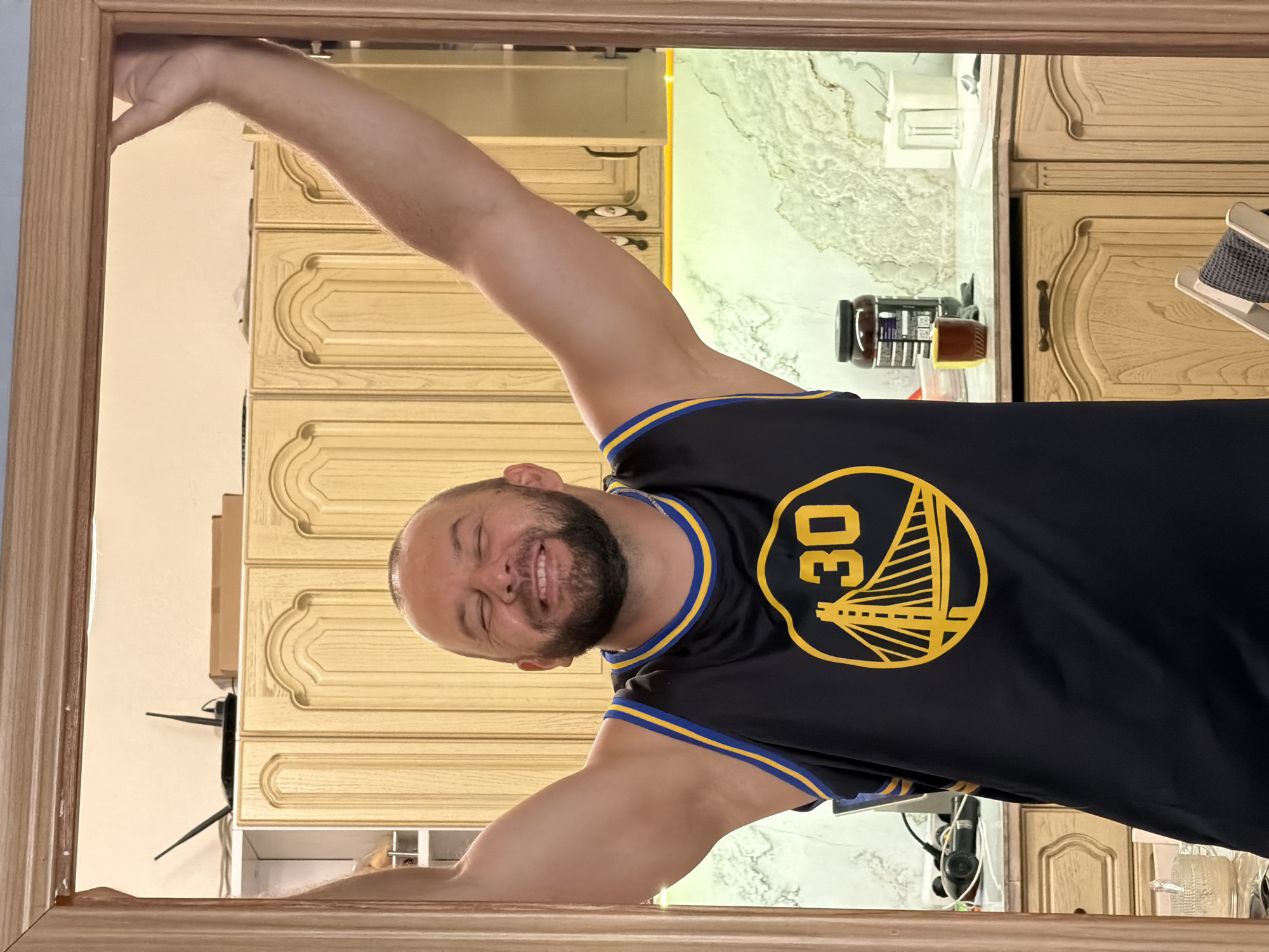 A man wearing a dark blue Golden State Warriors basketball jersey with yellow trim and the number 30 is standing with his arms raised, framing his head. He has a beard and is smiling with his eyes closed. He is positioned in a doorway, with a wooden frame visible on the sides. In the background, there are light-colored wooden kitchen cabinets. The countertop is a light-colored marble pattern, and a few objects are visible, including a dark-colored jar with a label, a small brown mug, and a white box. The lighting suggests an indoor setting, possibly a kitchen or dining area. The image does not provide any explicit information about the city of Sochi, Russia, or any specific location within it.