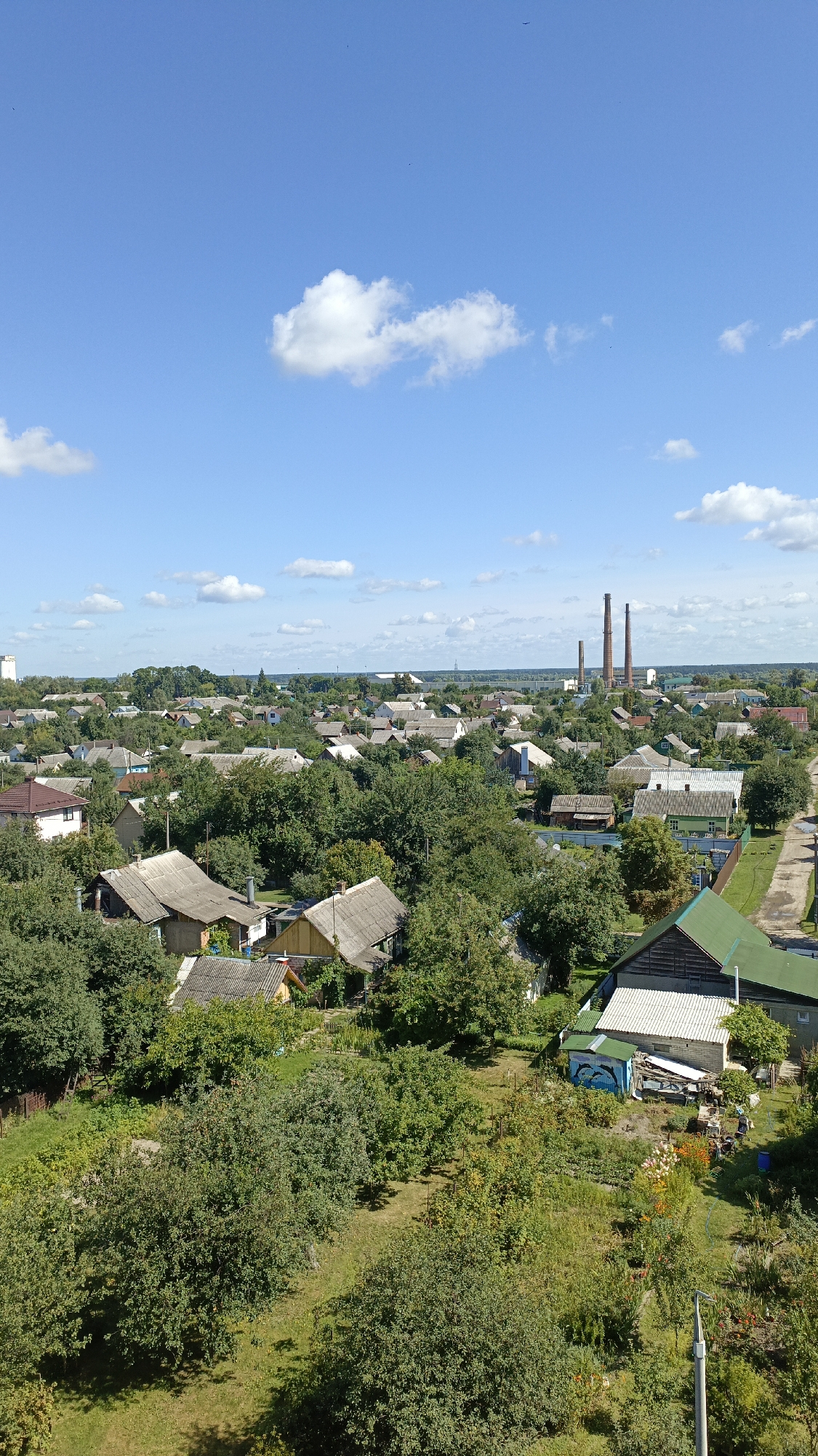 The image shows an aerial view of a residential area in Rechytsa, Belarus. The scene is dominated by numerous houses with pitched roofs, interspersed with lush green trees and vegetation. In the background, two tall industrial chimneys are visible against a bright blue sky with scattered white clouds. The houses appear to be single-family dwellings, many with surrounding gardens and yards. The overall impression is one of a rural or suburban setting.