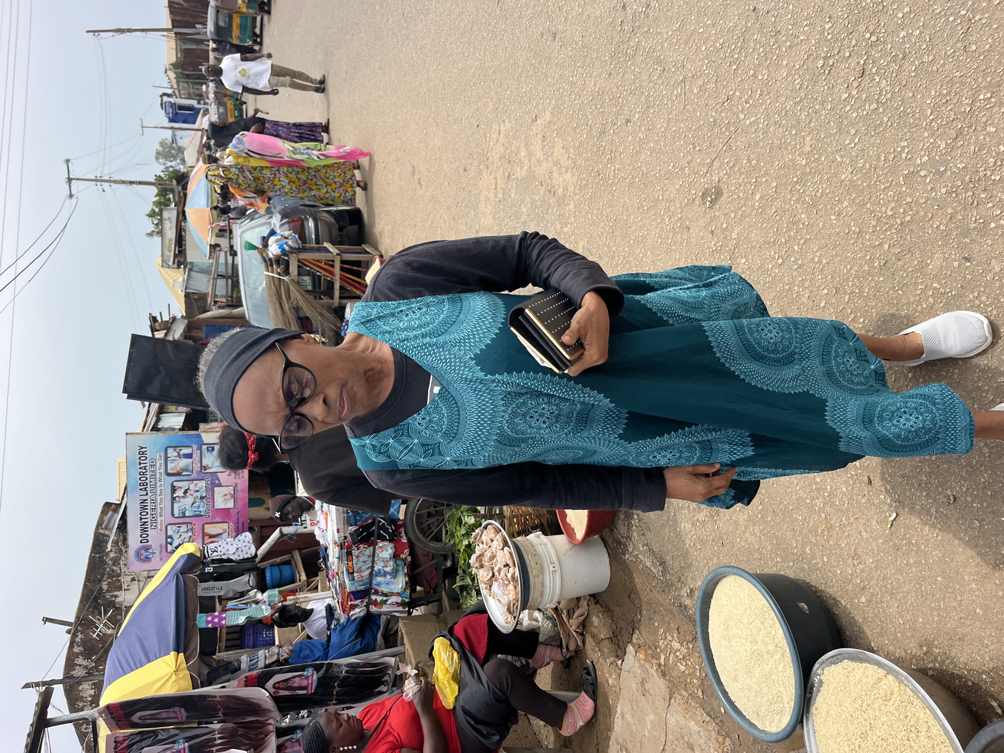 This media file captures a street scene in what appears to be a marketplace, possibly in Nigeria given the visible text. The primary subject is a woman wearing glasses, a black headscarf, and a teal-colored dress with an intricate pattern. She is holding a small black clutch with white dots. Her expression is neutral, and she is looking slightly downwards.
In the background, other people are visible, some engaged in activities related to commerce. There are stalls displaying various goods, including what looks like clothing, hair extensions, and food items. One stall has a sign that reads "DOWNTOWN LABORATORY NIGERIA LIMITED," with additional text about checking quality lab tests. Another stall has a colorful umbrella, and there are several large bowls filled with what appears to be rice. A few vehicles, including what look like auto-rickshaws, are also present in the distance, suggesting a busy thoroughfare.
The scene appears to be set during the daytime, with bright lighting, indicating fair weather. The ground is paved with rough asphalt. The overall impression is one of a bustling local market where people are shopping, selling, and going about their daily business.