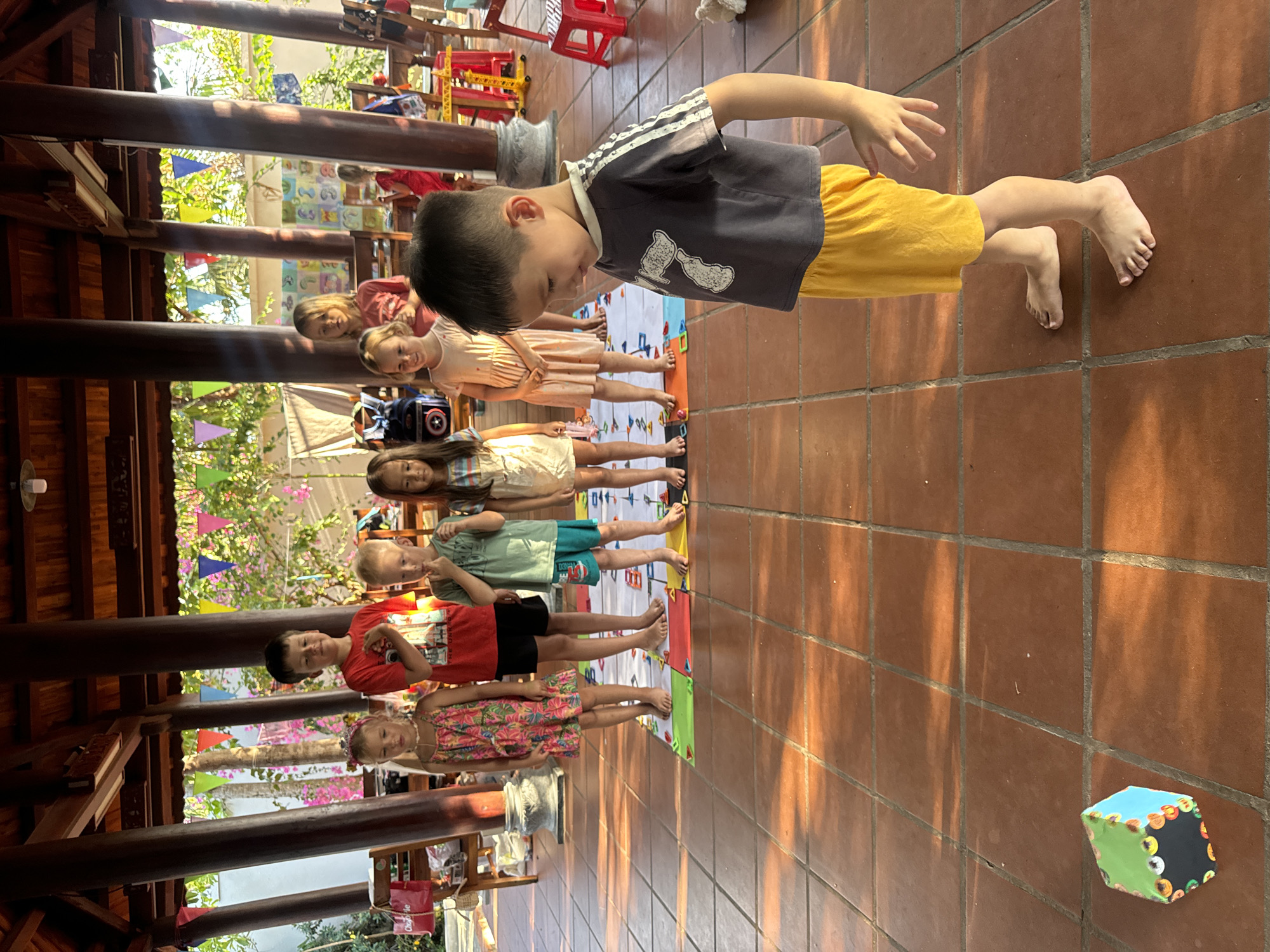 The image displays a line of approximately seven children, boys and girls, engaged in an activity in what appears to be an outdoor or semi-outdoor educational setting. The children, most of whom are barefoot, are standing on colorful mats or strips of paper laid out on a tiled floor.
In the foreground, a young boy wearing a dark grey t-shirt with white designs and yellow shorts is leaning forward, looking down with focused attention, suggesting he is interacting with the mat or an instruction. Behind him, other children are lined up, dressed in various casual attire including dresses, t-shirts, and shorts.
The setting features a reddish-brown tiled floor and a sturdy wooden beam structure overhead, indicative of a covered patio or open-air building. Bright natural sunlight illuminates the scene, casting distinct shadows on the floor, suggesting it is late morning or early afternoon. In the background, colorful pennant banners are strung, and lush green trees and foliage are visible, adding to the outdoor ambiance. Red plastic chairs and other classroom-like furniture are also present in the distance.
On the tiled floor to the right, a multi-colored cube (blue and green with circular "eye" patterns) is visible, potentially a prop for the children's activity. The overall impression is one of a playful or learning activity taking place in a warm, sunny environment, aligning with the specified location context of Mũi Né, Vietnam. No discernible text is visible in the image.