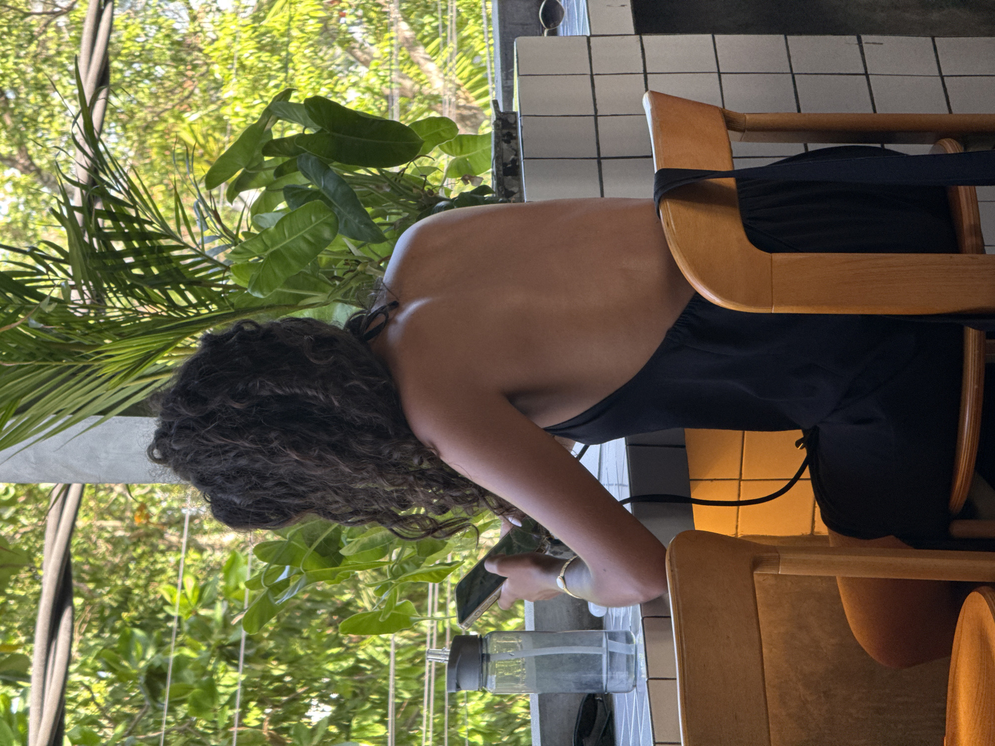 A woman with curly hair wearing a black dress is sitting on a light-colored wooden chair in what appears to be a cafe in North Kuta, Indonesia. She is holding a cell phone in her left hand and looking at it. Next to her is a water bottle on a table with white tile. Lush green vegetation is visible through a window behind her.
