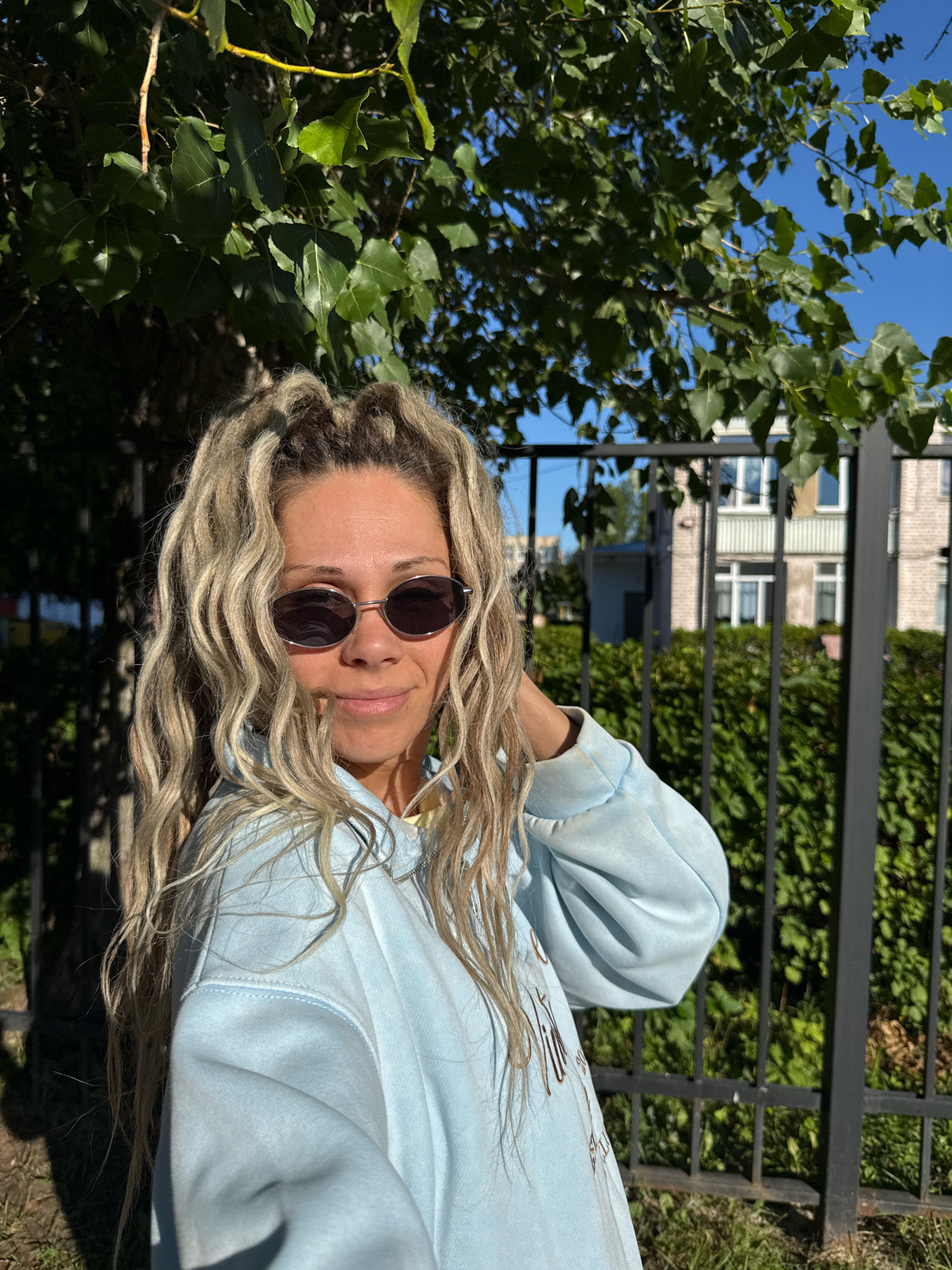 A blonde woman with dreadlocks is posing for a photo outside, in front of a black metal fence. She is wearing a light blue hooded sweatshirt and sunglasses. Behind her, there is a dense green hedge and a building with light-colored walls and dark windows. Sunlight is casting shadows from tree leaves on the left side of the image. The scene appears to be in a residential area.