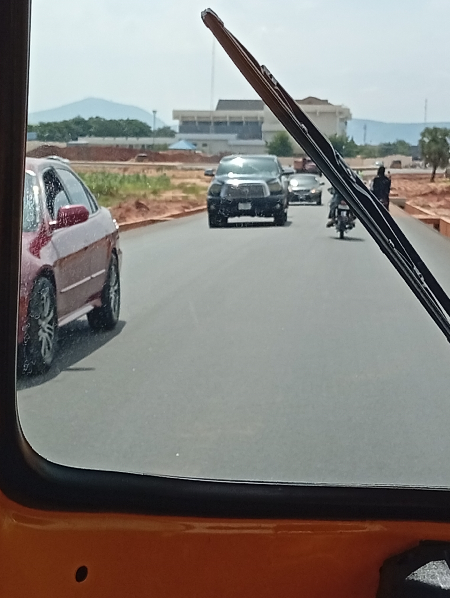 The image is a street-level view from inside a vehicle, looking forward through a windshield. The scene depicts a road in the city of Gombe, Nigeria, with several vehicles and a person on a motorcycle.
In the foreground, the orange body of the vehicle from which the photo is taken is visible at the bottom. A windshield wiper partially obstructs the view.
On the road, a dark-colored SUV is in the center lane. To the left of the SUV, a red car is visible, with its rear tire showing. Further ahead, a motorcycle with a rider is in the right lane. To the right of the motorcycle, a person wearing dark clothing is standing near the side of the road.
In the background, a large building with blue seating, possibly a stadium or arena, is visible. Beyond that, a distant mountain range can be seen under a hazy sky. The surrounding landscape appears to be arid with patches of greenery and construction sites. The road surface is asphalt. The time of day appears to be daytime, with bright, diffused sunlight.