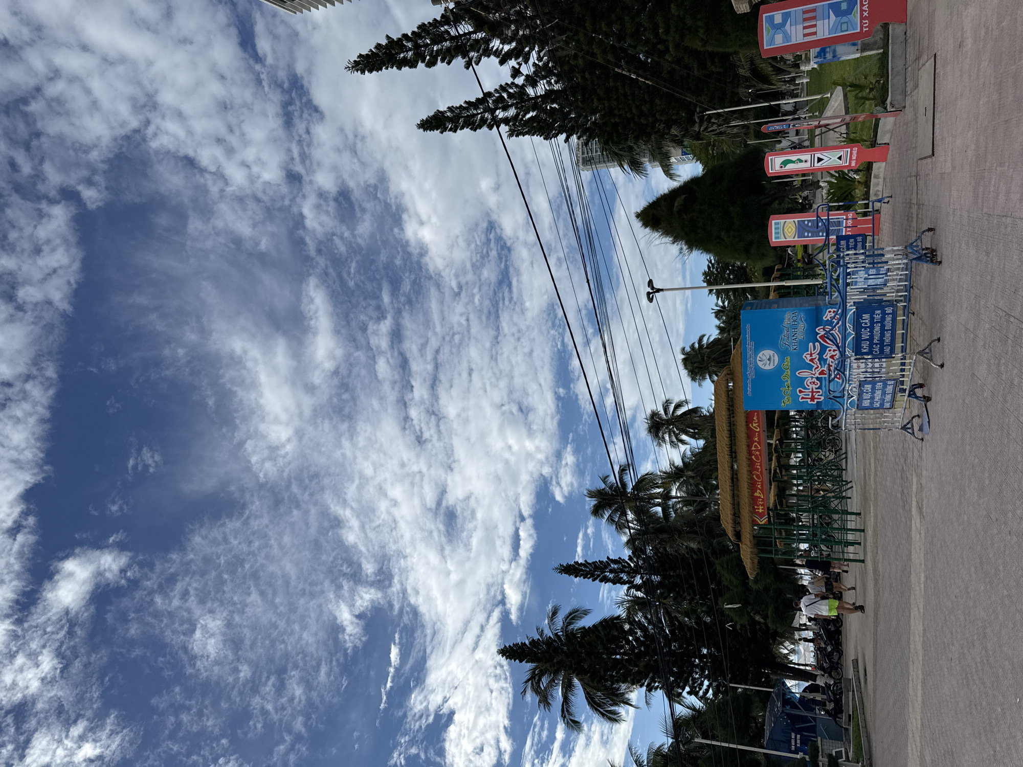 This image depicts a street scene in Nha Trang, Vietnam, under a bright blue sky with scattered white clouds. In the foreground, a paved walkway is lined with metal barriers, one of which is blue and white. Several colorful, upright signs with abstract designs and Vietnamese text are visible on the right side of the walkway. Further back, a cluster of bicycles is parked near a structure with a thatched roof supported by green metal poles. To the left, a blue kiosk-like building with text and a motorcycle are present. Palm trees and tall, dark green coniferous trees are interspersed throughout the scene, with a modern building visible in the background on the right. Three individuals are walking away from the camera on the paved area, one wearing a white t-shirt and yellow shorts, another in a white t-shirt and dark pants, and the third in a dark top and pants. The overall environment suggests a public area, possibly near a tourist attraction or park in Nha Trang.