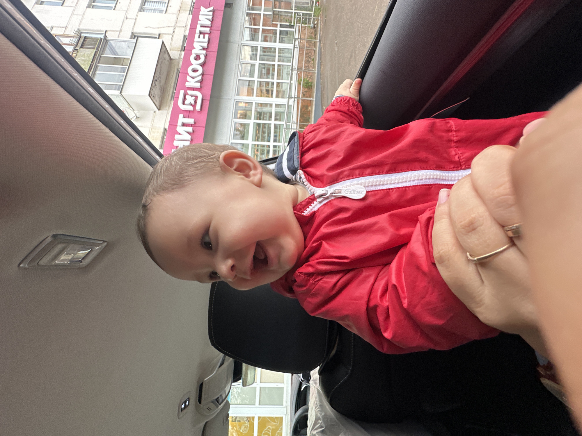 An infant, wearing a red jacket, sits in the back seat of a car, looking out the window. The infant has light skin and is smiling. In the background, a building with a large pink sign reading "ШИТ К КОСМЕТИК" is visible. The setting appears to be an urban environment.