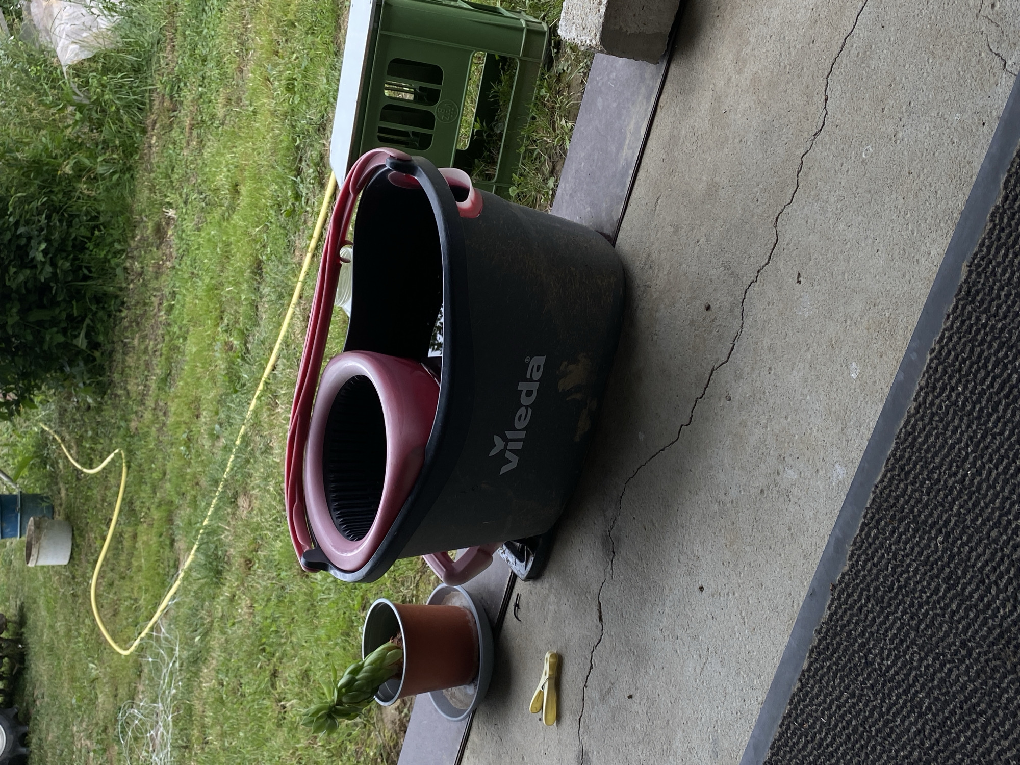 An outdoor scene shows a dark gray Vileda brand mop bucket with a red rim and handle, sitting on a concrete patio. Inside the bucket, there is a pink mop spinner. To the left of the bucket, a small potted plant with green leaves sits on a gray saucer, next to a yellow clothespin. In the background, a grassy area is visible with a yellow hose laid out on the grass and a green plastic crate. The environment appears to be a yard or garden.