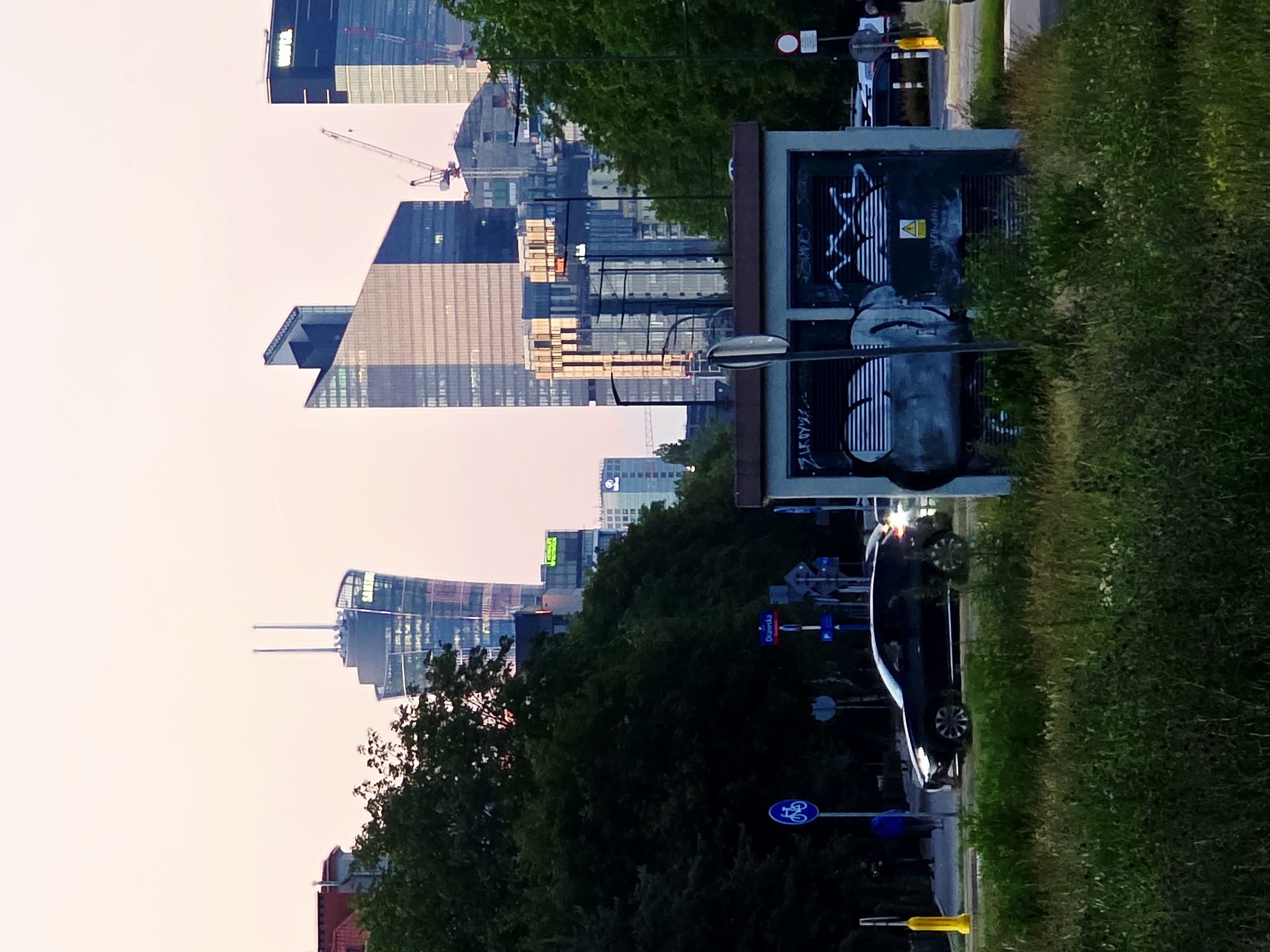 The image depicts a street scene in Warsaw, Poland, at dusk. The sky is a soft pink and orange hue, indicative of twilight. In the background, a cluster of modern skyscrapers dominates the skyline. One prominent building, possibly the Sky Tower or a similar structure, features a distinctive curved design and is topped with two spires. Other skyscrapers are visible, some with cranes nearby, suggesting ongoing construction.
In the foreground, a dark-colored SUV is driving on a paved road. A pedestrian walkway is visible to the left, with a blue sign indicating a bicycle path. Several streetlights and traffic signs are present. A small, utilitarian building, likely an electrical substation, stands in the mid-ground and is covered in graffiti. Dense green trees line the street, partially obscuring the view of the buildings behind them. The ground in the immediate foreground is covered with tall, green grass and some wildflowers. The overall impression is of a developing urban environment where nature and modern architecture coexist.