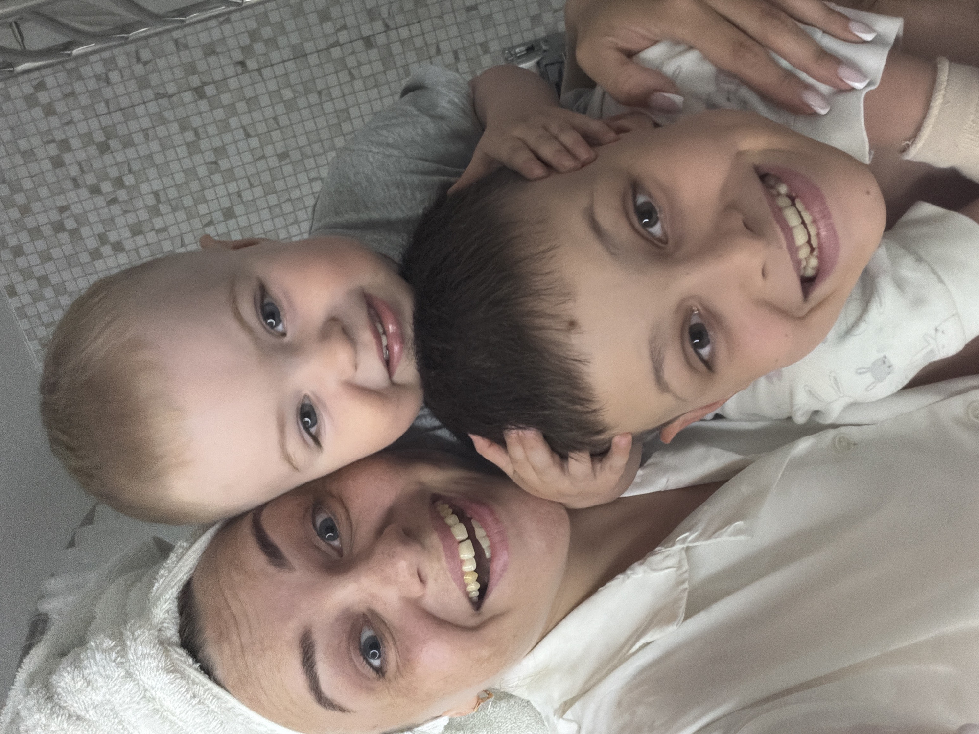 A woman and two children are lying on a tiled surface. The woman is wearing a white towel on her head and a white button-down shirt. The younger child has blond hair and is wearing a gray shirt. The older child has brown hair and is wearing a white shirt with a small rabbit print. The children are touching the woman's face. They are all smiling. The background appears to be a bathroom with small, square tiles. The location is unknown.
