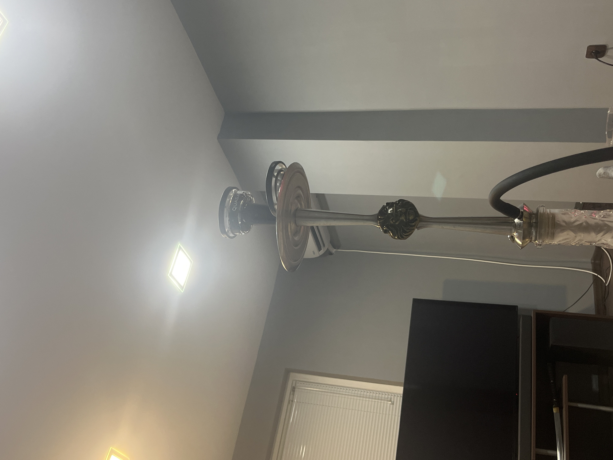 A hookah stands in the center of the frame, reaching towards the ceiling. The hookah has a metallic finish and features an ornate design near its base, resembling a lion's head. A black hose curves away from the hookah towards the right of the image. In the background, a large, dark television screen is visible on the left, mounted above a dark entertainment stand. A window with horizontal blinds is situated to the left of the television. The walls are painted a neutral grey. Two square ceiling lights illuminate the room, one with a warm, yellowish glow and the other with a bright white light. The scene appears to be indoors, in a room within Montana, Bulgaria.