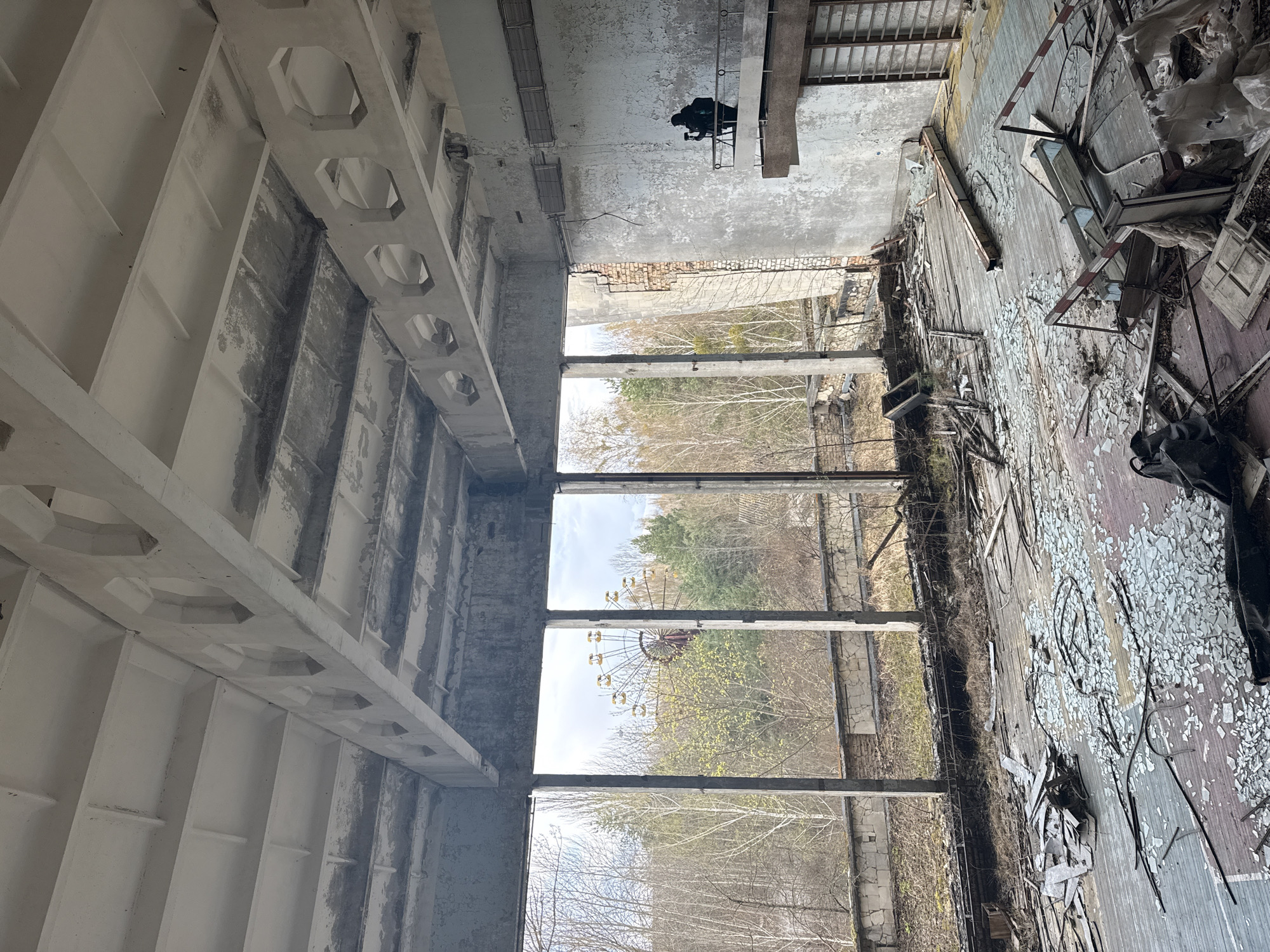 The image shows an interior view of an abandoned building in Pripyat, Ukraine, with a deteriorated basketball court floor covered in debris and shattered glass. Large windows offer a view of a Ferris wheel, a recognizable landmark of the abandoned city. A person is visible near the ceiling, possibly a photographer. The building's concrete structure shows signs of decay.