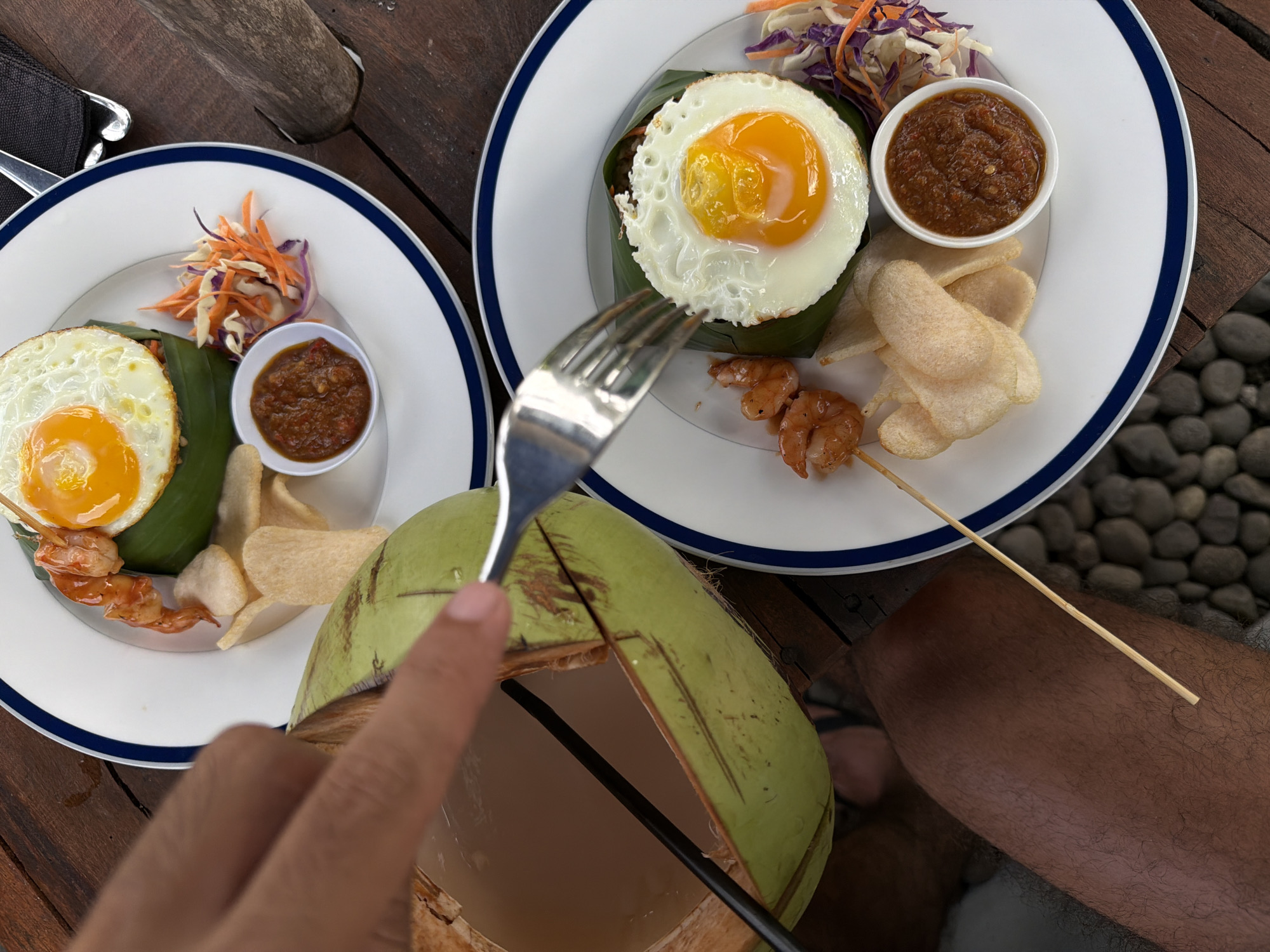 The image shows a partially obscured view of two plates of food and a coconut. The plates are white with a blue rim and contain a fried egg on a green leaf, a small bowl of a reddish sauce, a pile of shredded vegetables, and prawn skewers. One plate also has white, puffy snacks. A hand holding a fork is visible in the foreground, near a green coconut that has been cut open and has a black straw inside. The scene is set on a wooden surface, likely a table. In the background, there are small grey rocks. The location is Selemadeg Timur, Indonesia.