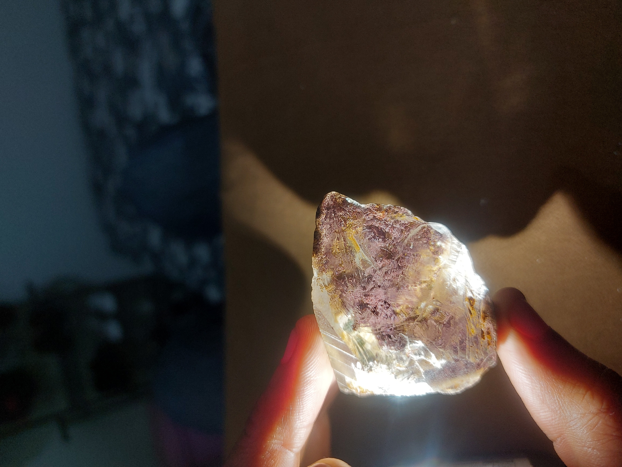A person is holding a raw, translucent amethyst crystal up to the light. The crystal shows a mix of purple, brown, and clear hues. The background is dimly lit with mixed textures of dark and light patches. The image location within Dar es-Salaam, Tanzania, cannot be determined from the visual information provided.